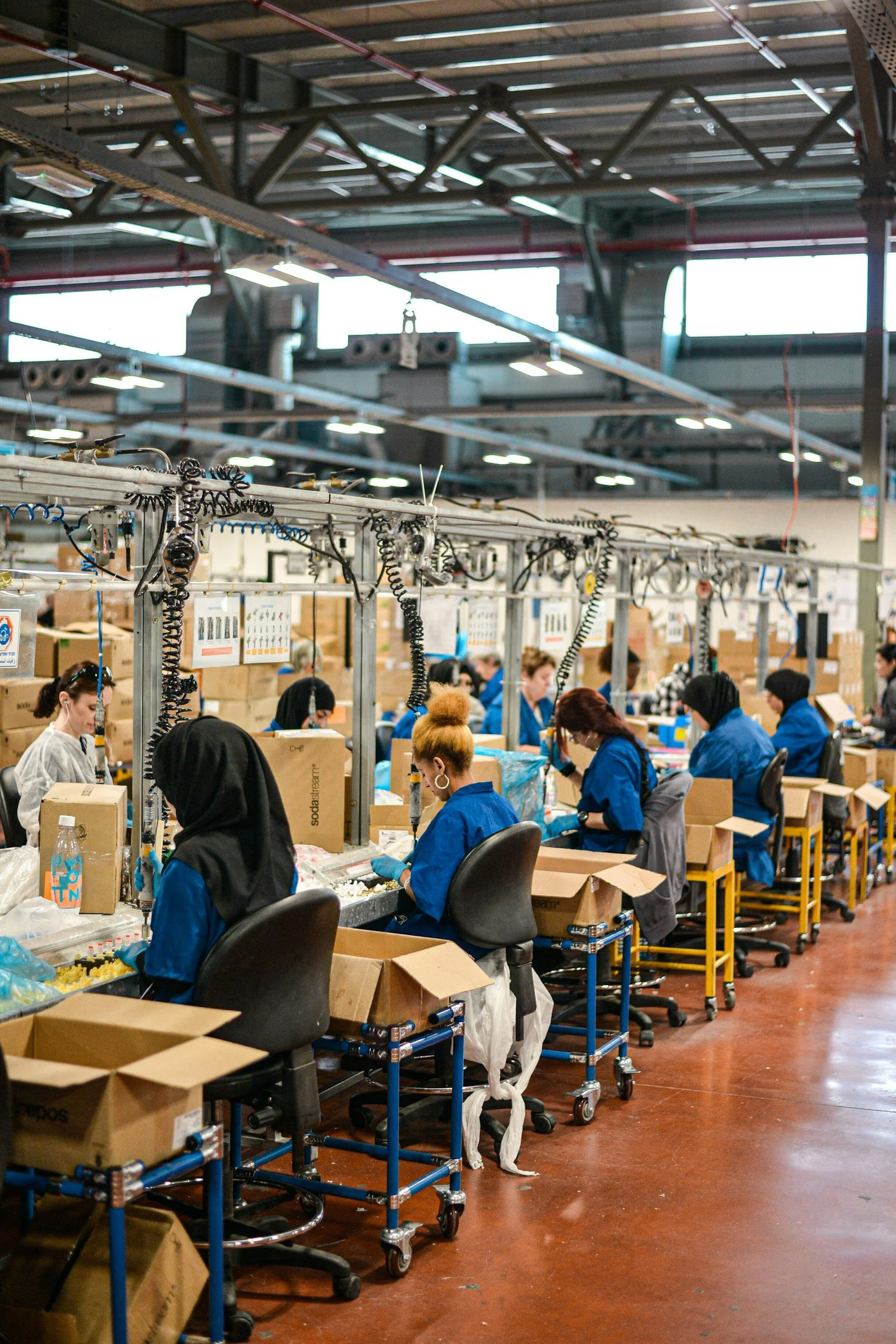 Factory workers seated at assembly stations packaging products in a large industrial space with metal beams and bright lighting.