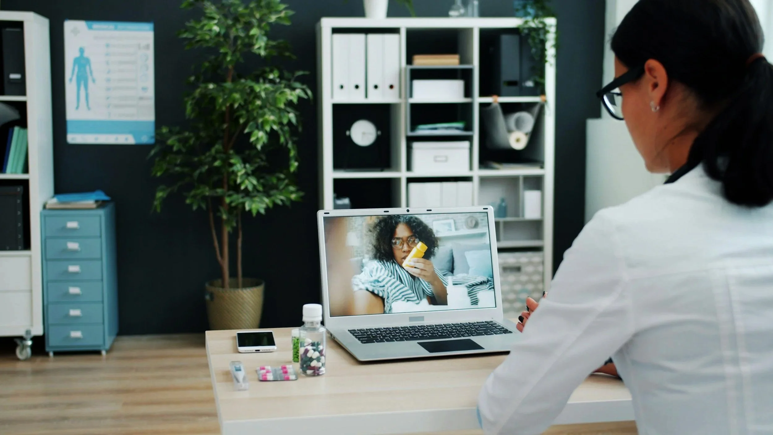 A healthcare professional wearing glasses and a white coat sitting at a desk, during a video consultation with a woman on a laptop screen. The woman appears to be taking pills and looks surprised. The desk has medication bottles and a smartphone, and the room has shelves, a plant, and a poster on the wall.