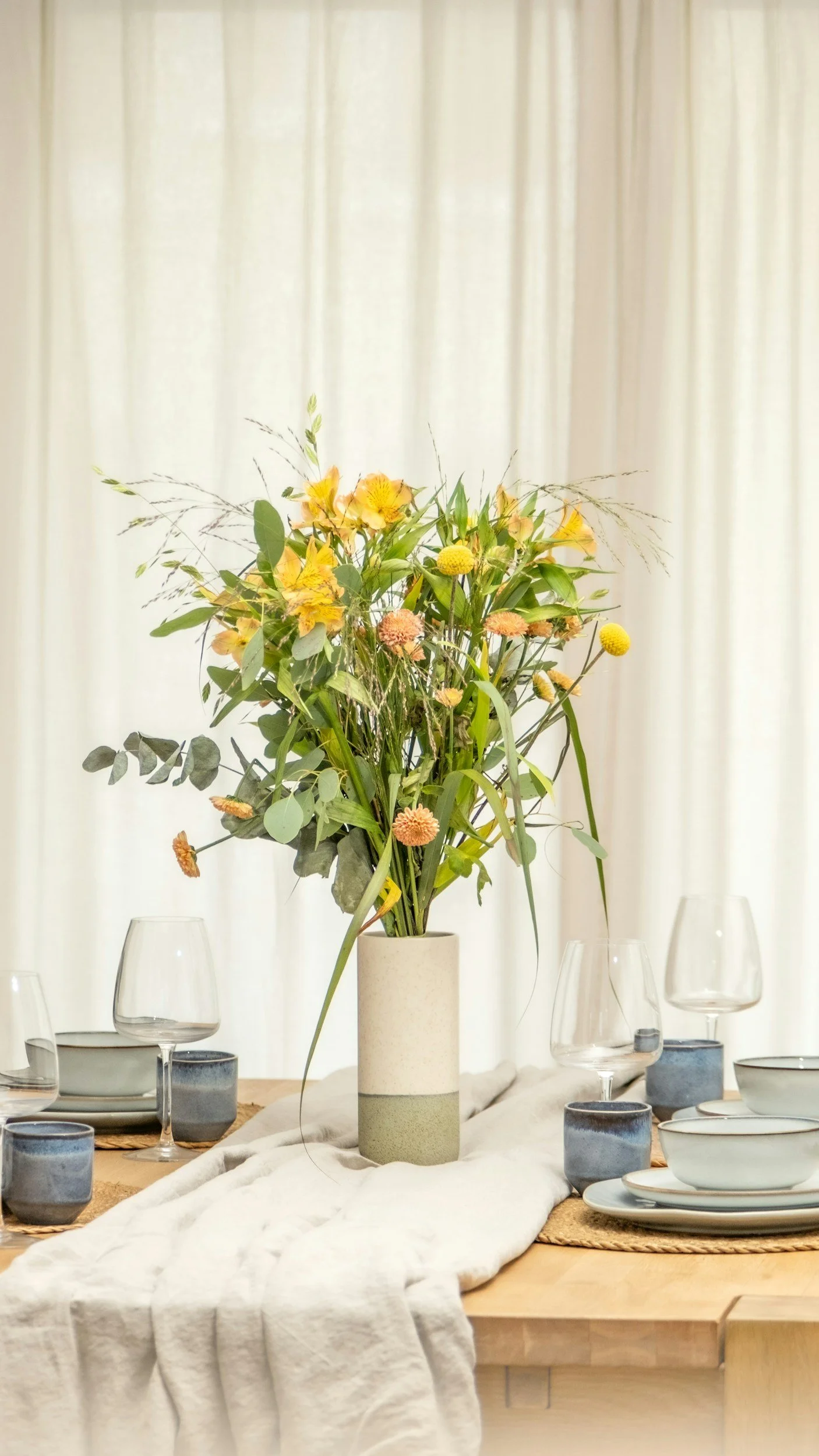 A tall beige and gray ceramic vase holding a large bouquet of yellow and orange flowers with green foliage, set on a wooden table covered with a white cloth, surrounded by empty glasses and plates, with a neutral curtain background.