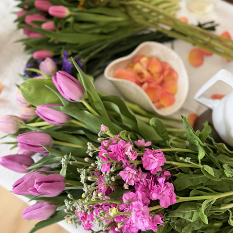 Assorted pink and purple flowers on a table with a white bowl of peach-colored flower petals nearby.
