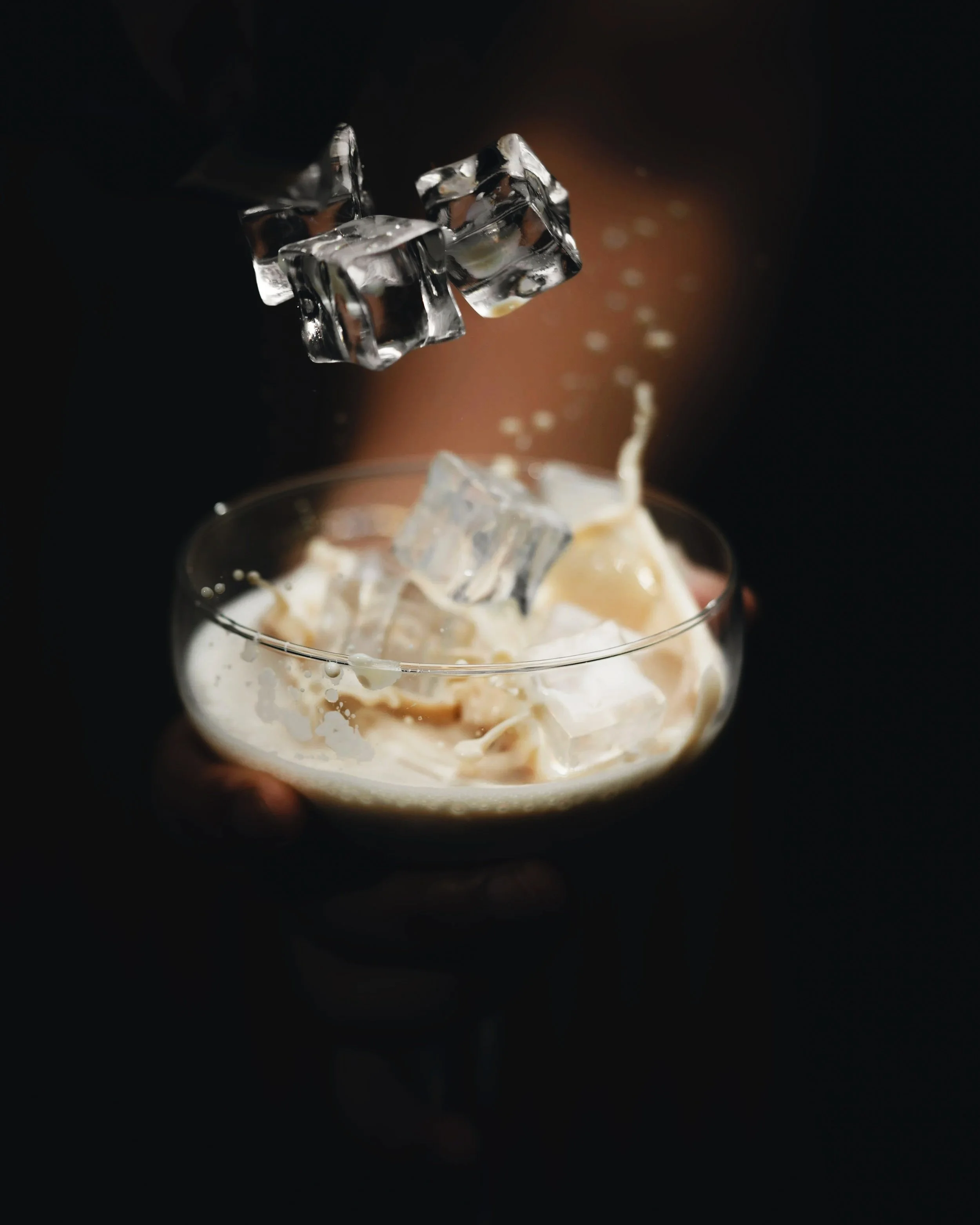 Ice cubes dropping into a creamy beverage in a glass, against a dark background.