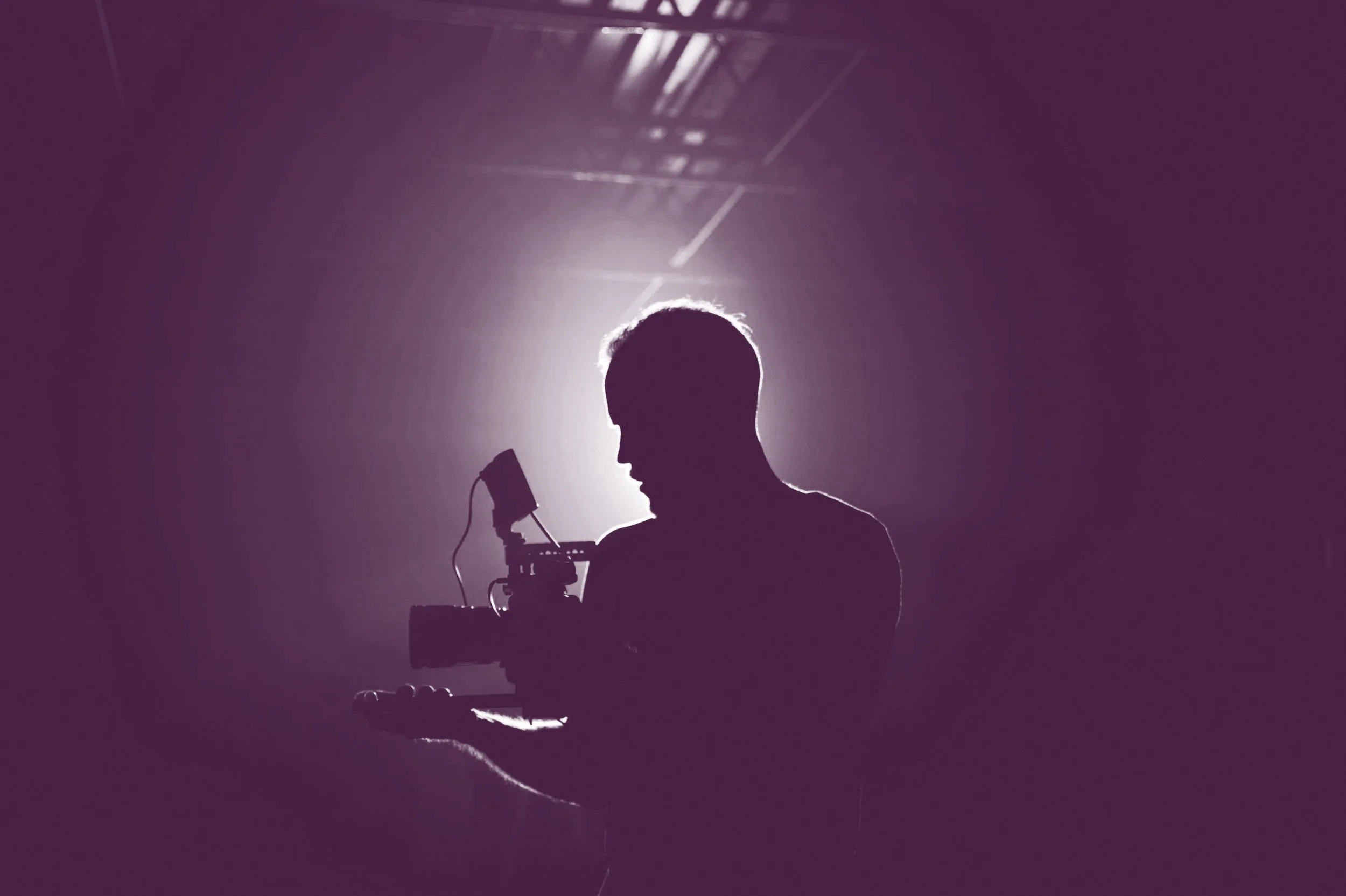 Silhouette of a person holding a camera, backlit by bright light, with a dark purple background.