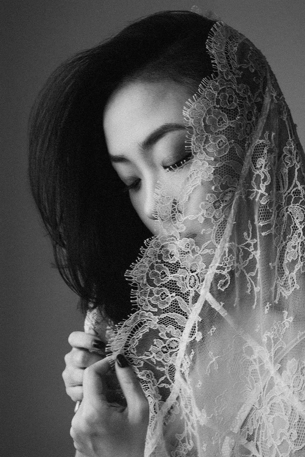 A black and white portrait of a woman with closed eyes, holding lace fabric near her face.