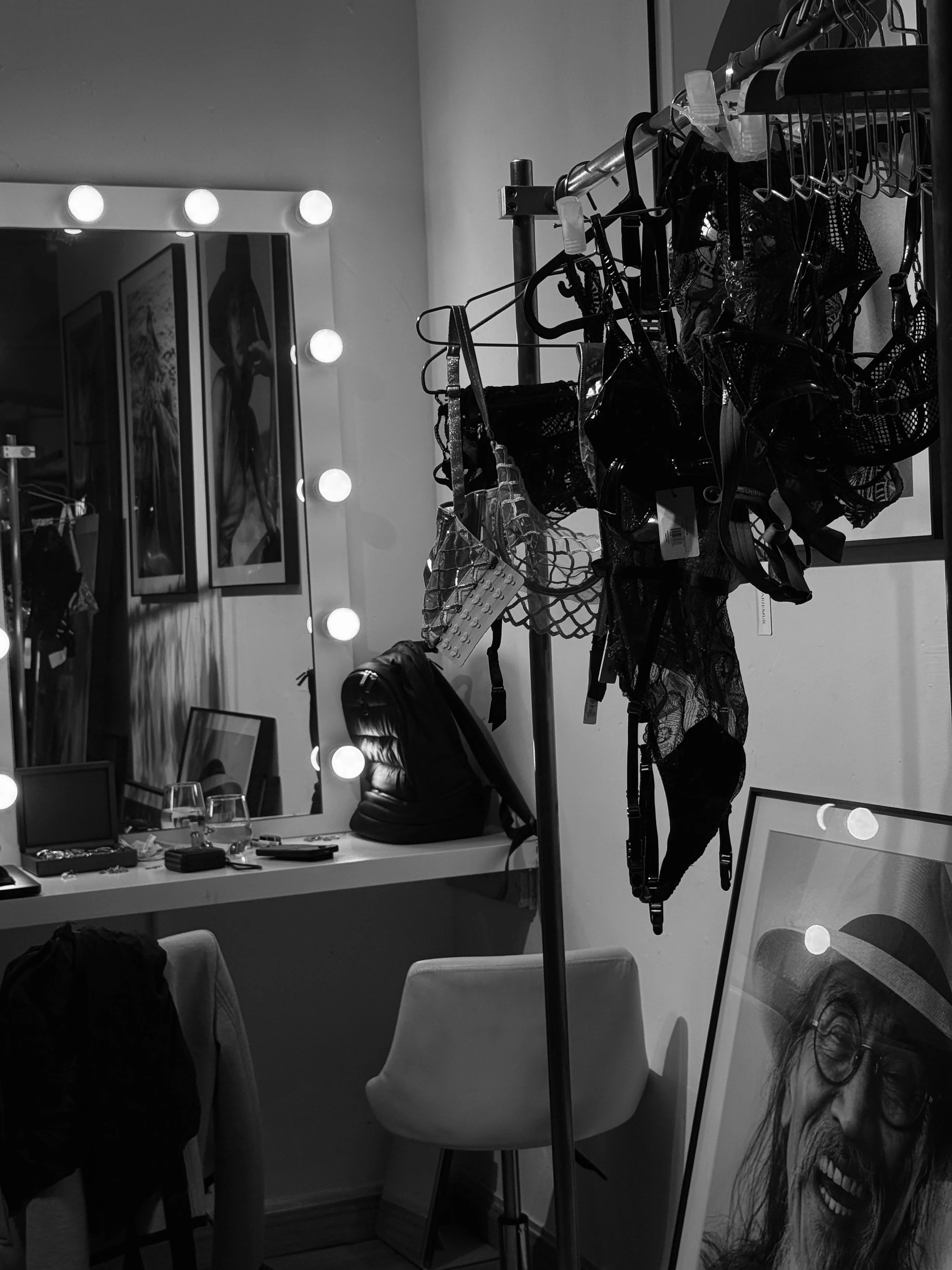 A black and white photo of a dressing room with a large mirror surrounded by light bulbs, a table with jewelry and other items, a chair, a clothing rack with various lingerie or clothing items hanging, and a framed portrait of an older man wearing glasses and a hat, grinning.