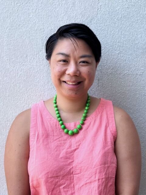 Person wearing a pink sleeveless top and green bead necklace against a light wall background.