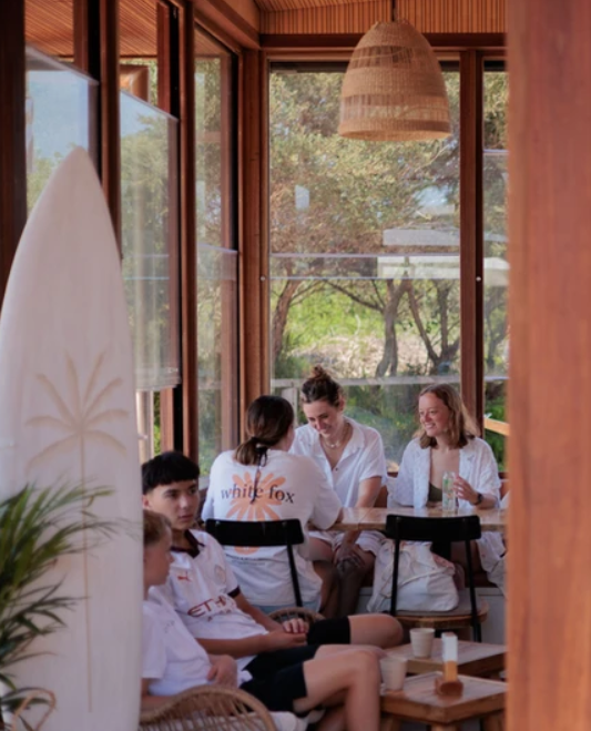 A group of children and a woman sitting around a table inside a wooden sunroom with large windows, enjoying a meal or drinks on a sunny day.