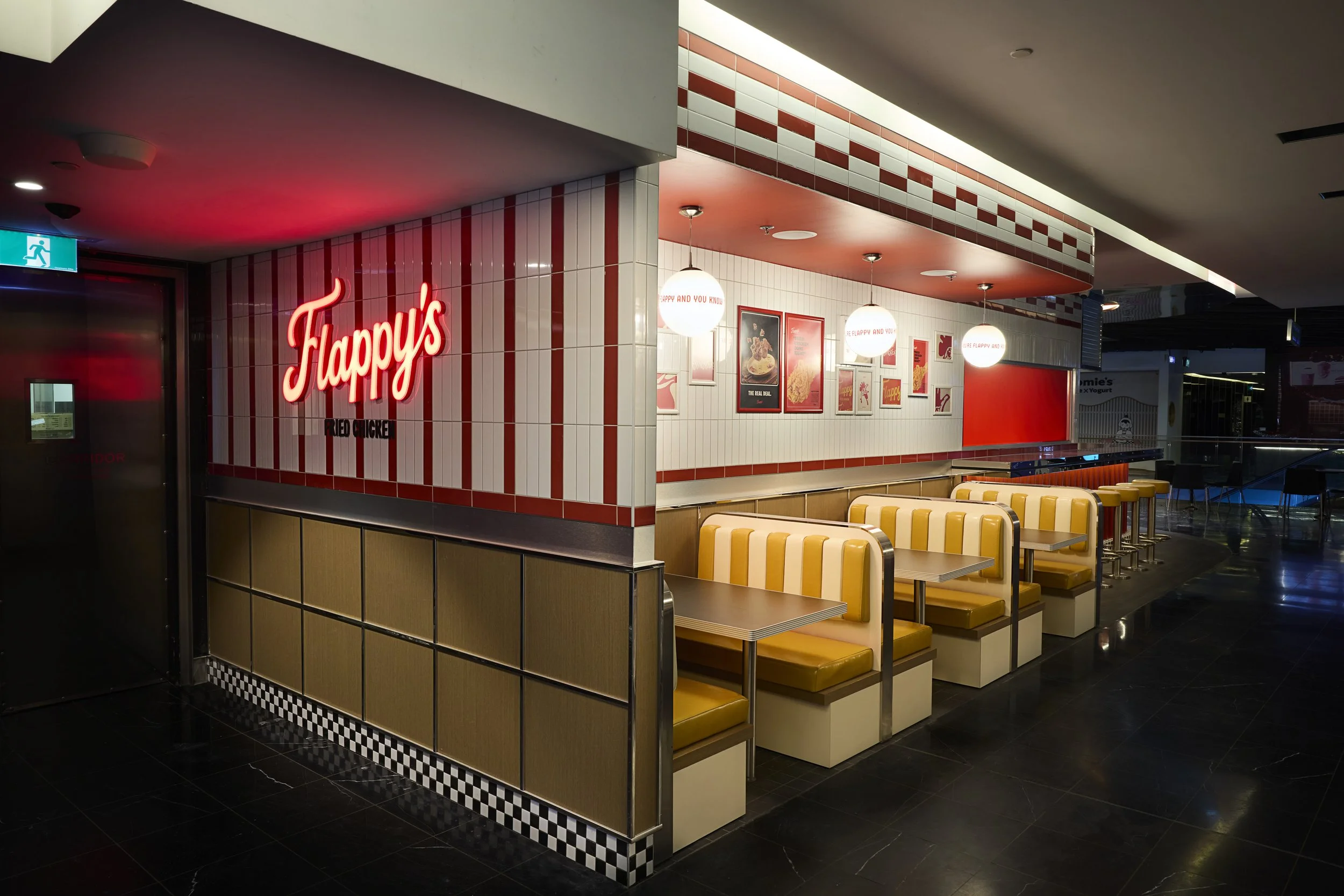 Interior of a fast-food restaurant named 'Flappy's' with yellow and white striped booths, framed menu posters on white tiled walls, hanging globe lights, and a neon sign reading 'Flappy's' in red.
