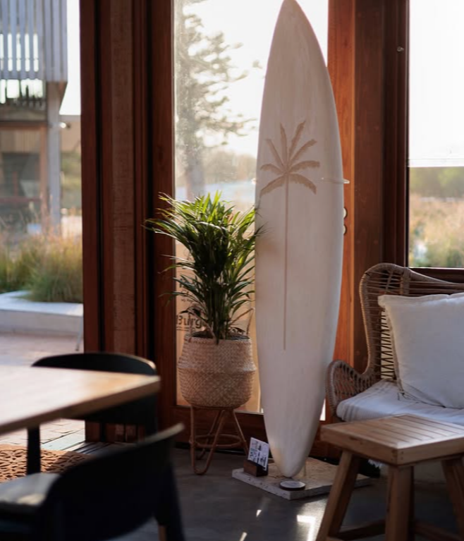 A surfboard with a palm tree design leaning against a wooden wall next to a potted plant and a wicker chair with a cushion, inside a room with large windows.