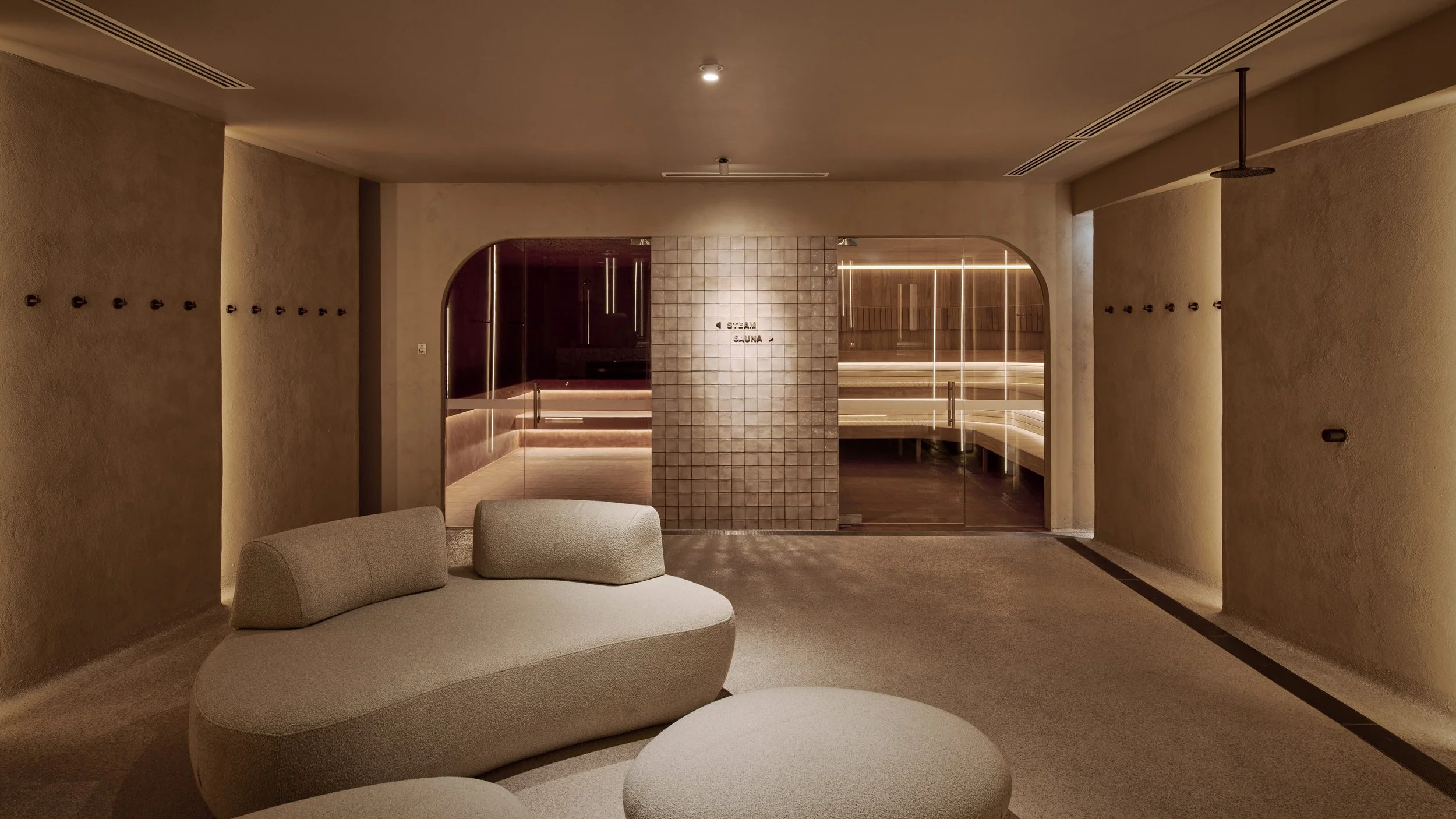 Modern spa lounge area with beige sofa and soft lighting, leading to a sauna room with glass doors and tiled interior.