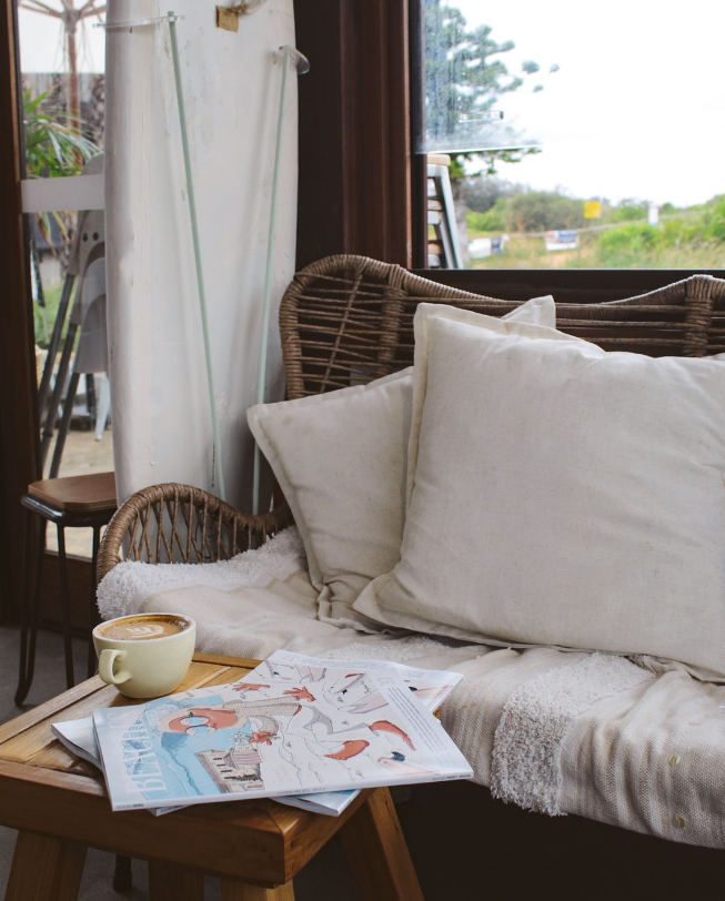 A cozy indoor scene with a wicker sofa, beige pillows, a wooden table with a cup of coffee and magazines, and a large window showing an outdoor view with trees and a cloudy sky.