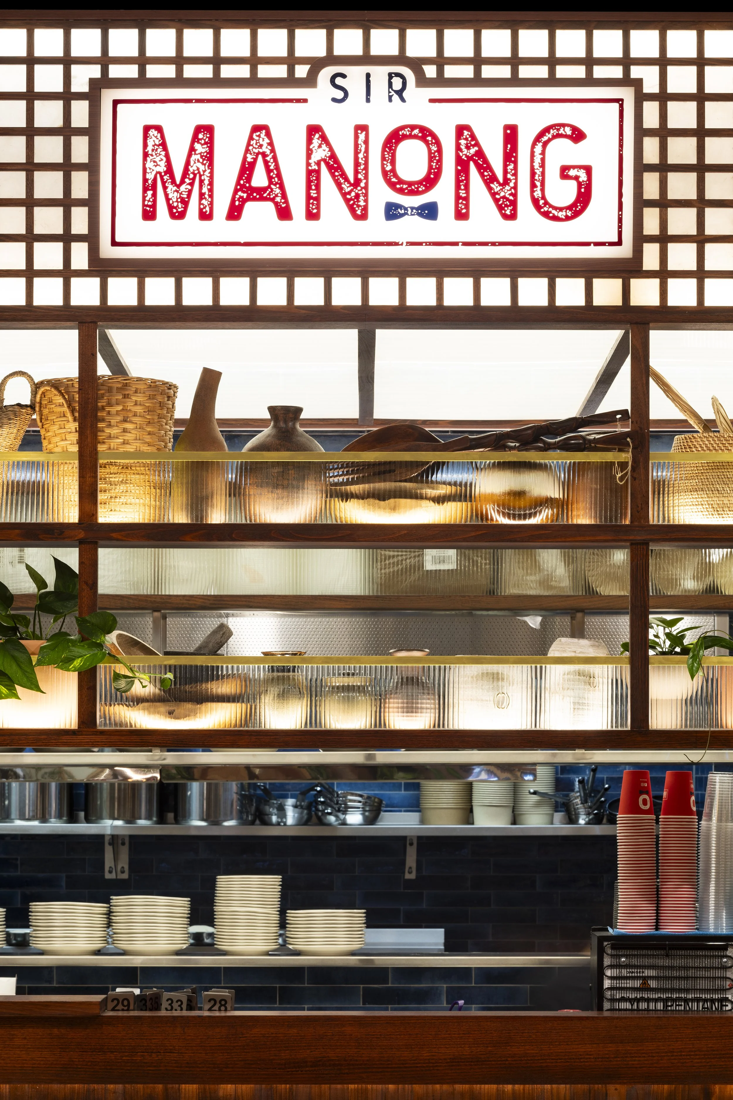 A restaurant order counter with a large illuminated sign that reads 'Sir Manong'. The backdrop includes shelves with woven baskets, pottery, and kitchen tools, with stacked plates and cups below.