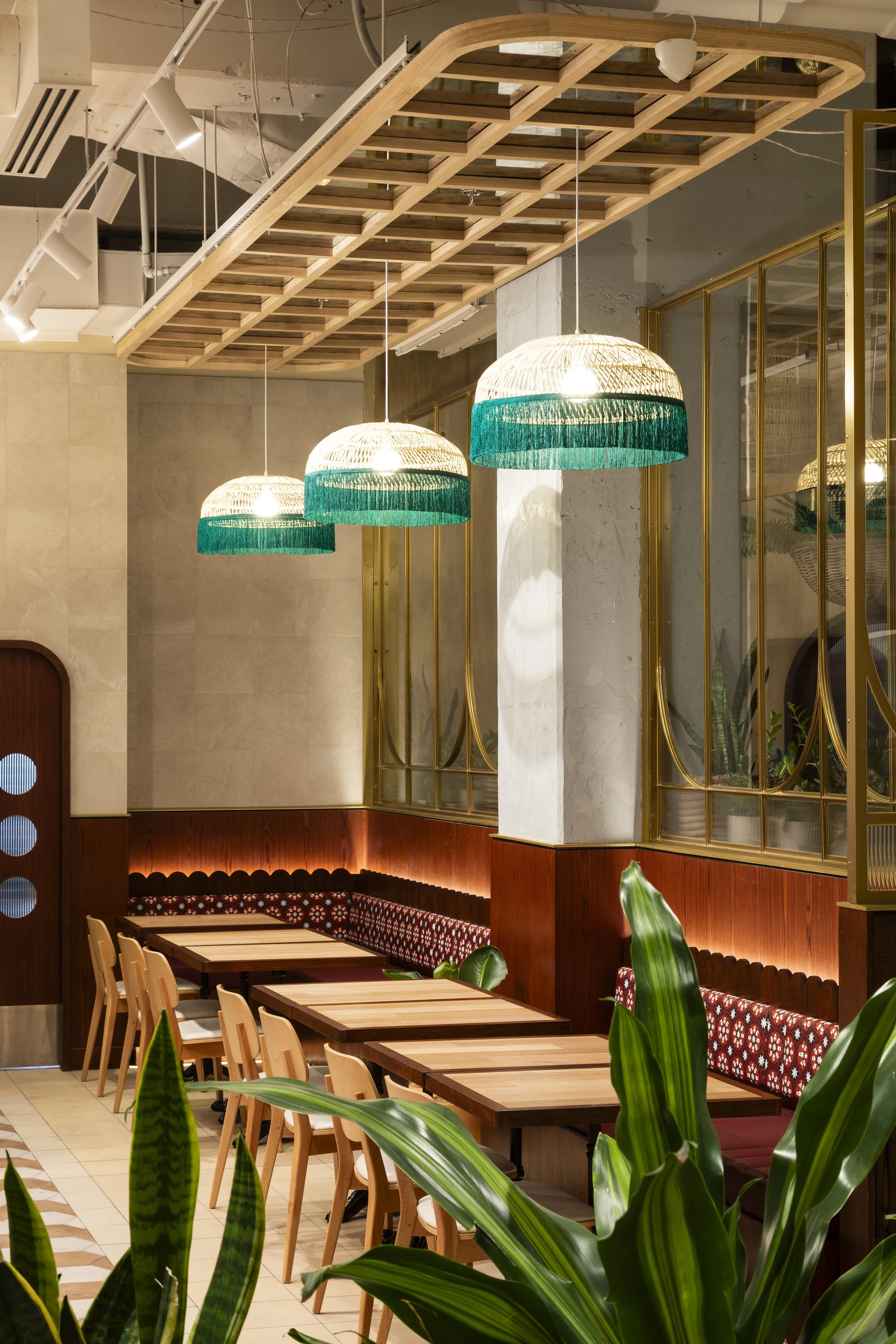Interior of a restaurant with wooden tables, chairs, and cushioned bench seating against the wall. Three pendant lights with woven shades and green trim hang above. Green plants are in the foreground, and gold-framed glass panels separate sections of the space.