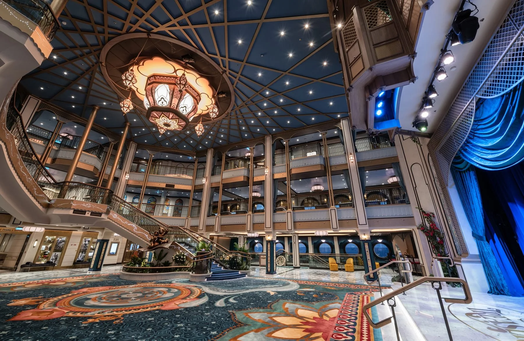 Interior of an elegant multi-level hotel lobby with a large ornate carpet, a stairway, a sculpture of a person, a chandelier, blue curtains, and multiple floors with railings.