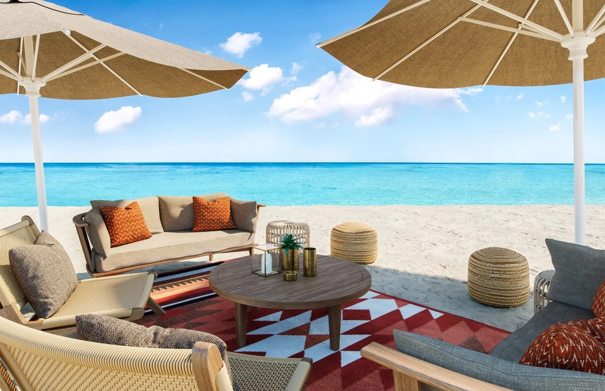 Outdoor beach lounge area with cushioned chairs, a sofa with pillows, a wooden coffee table with decorative items, woven stools, and large umbrellas, overlooking the ocean and a bright blue sky with clouds.