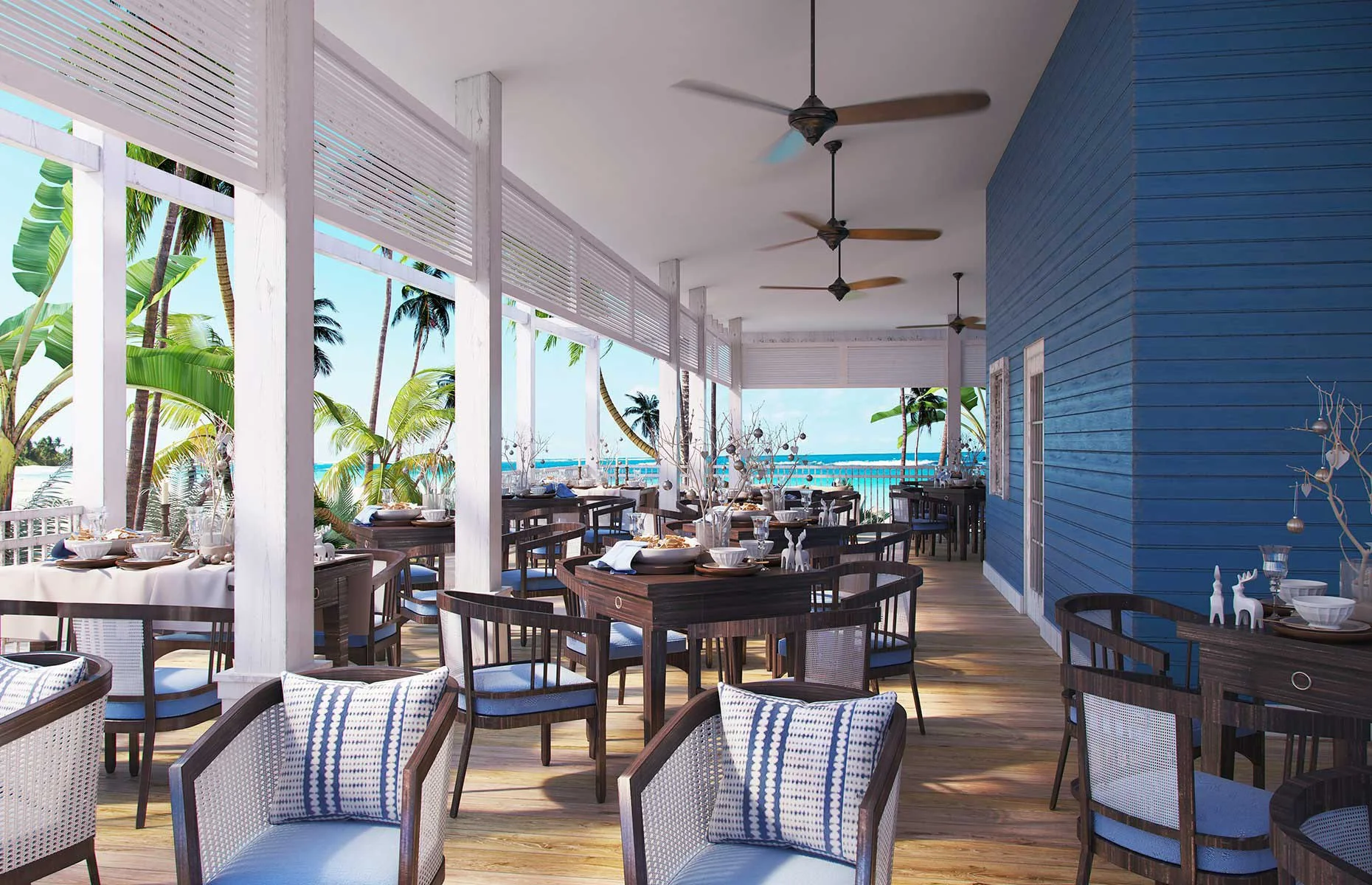 A bright seaside restaurant with wooden furniture, decorated for a meal, overlooking the ocean on a sunny day, with palm trees visible outside.
