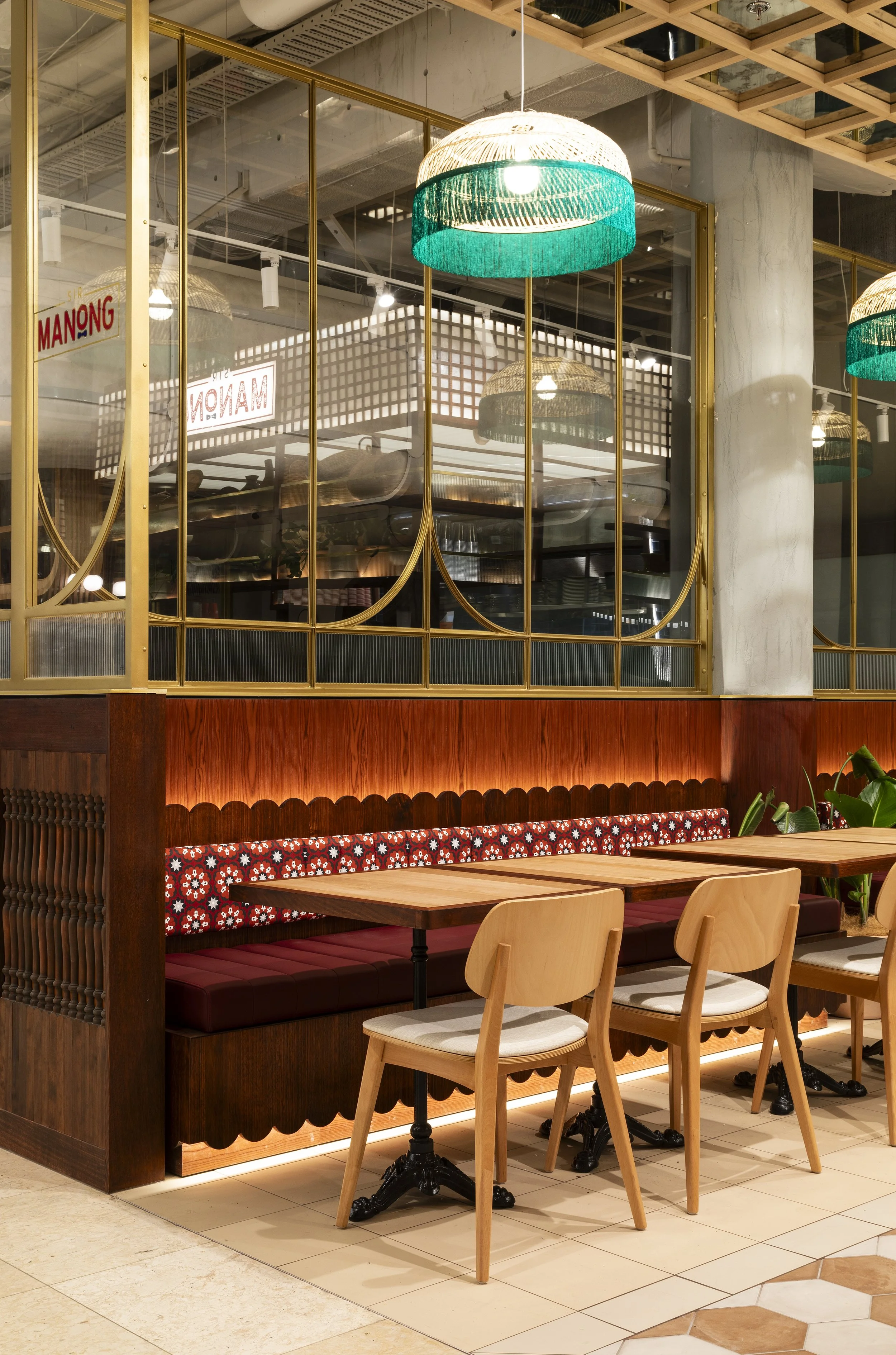 Indoor seating area with wooden tables and chairs, decorative wood paneling, and large hanging lamps with woven shades and green accents, separated by glass panels with gold frames.