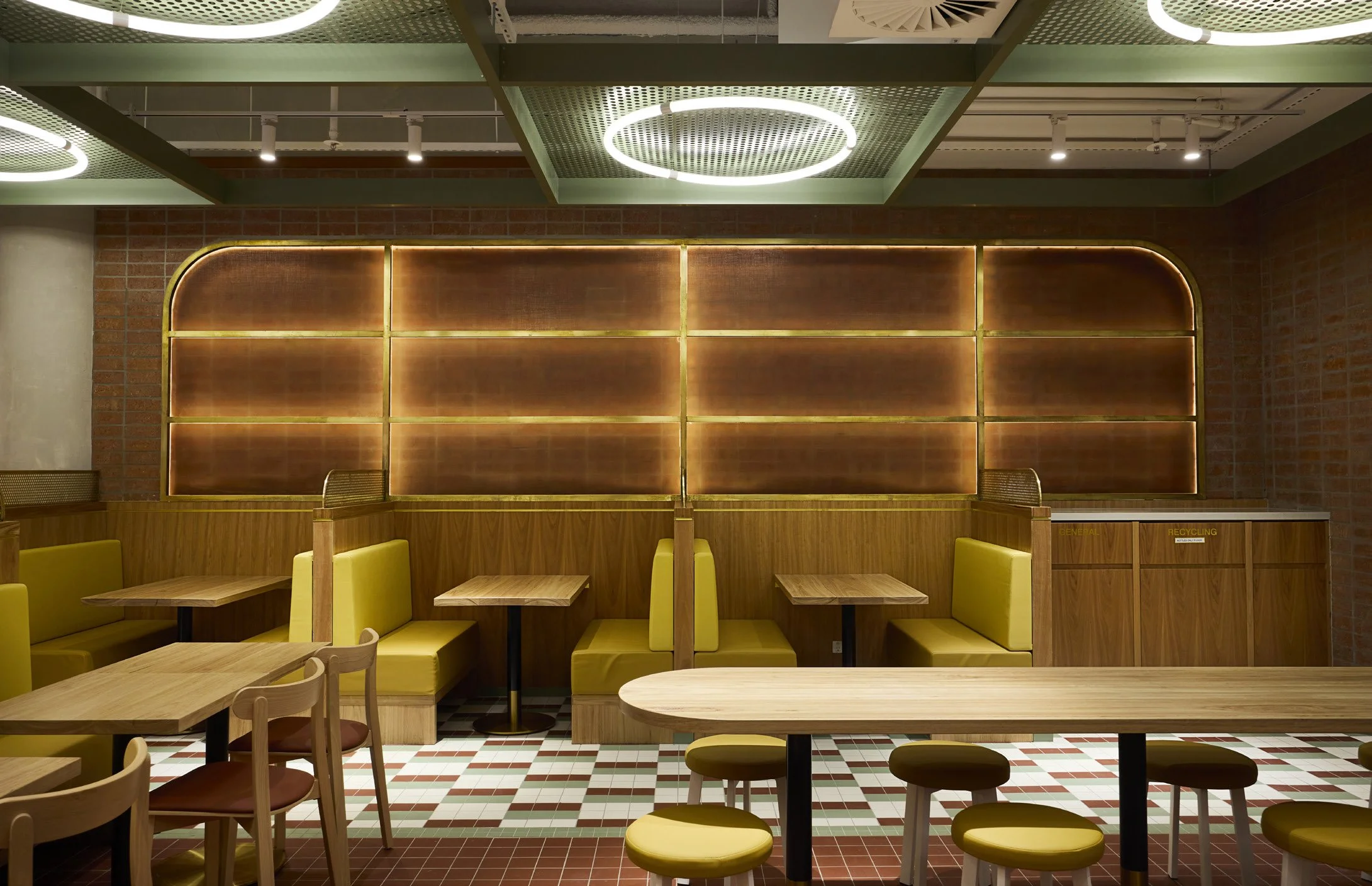 Interior of a modern restaurant with yellow seating booths, wooden tables, and a patterned tile floor, illuminated by ceiling lights and a decorative wall with backlit panels.