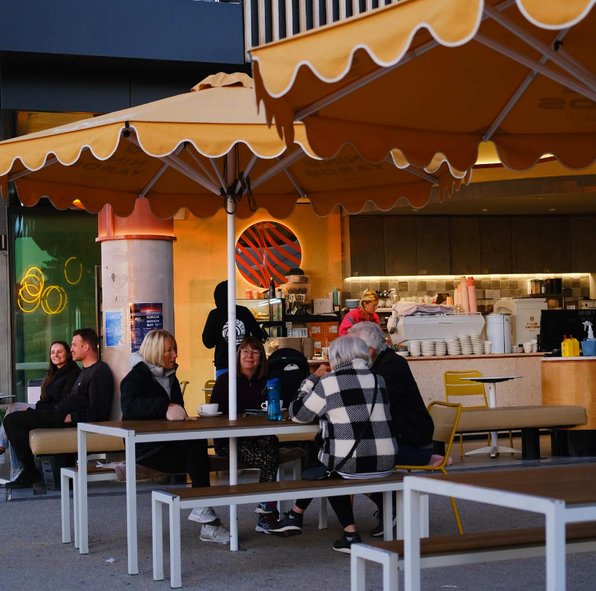 Outdoor cafe seating area with yellow umbrellas, several people sitting at tables, and a coffee stand in the background.