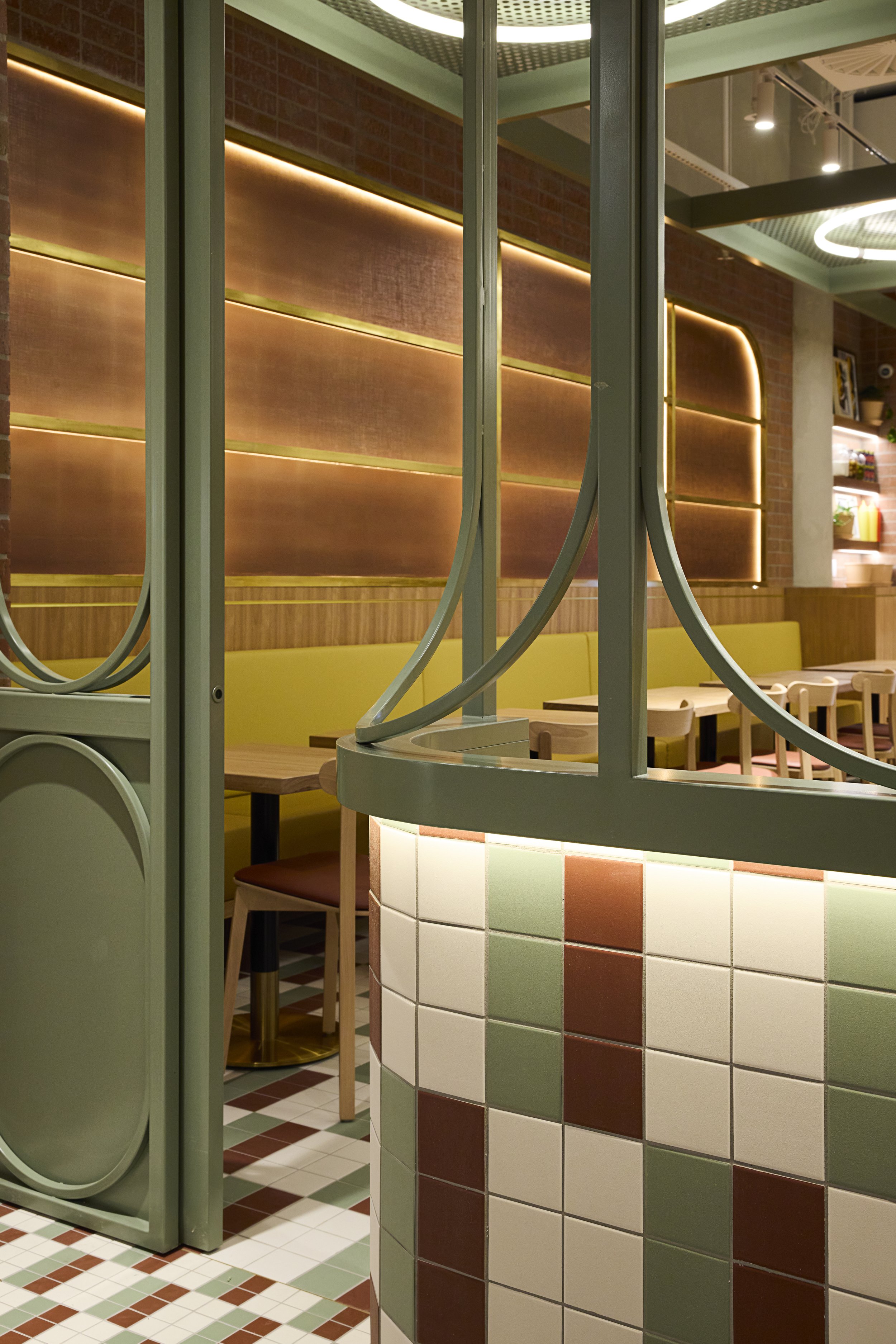 Interior view of a modern restaurant or cafe with a green and brown color scheme, featuring tiled and upholstered seats, wooden tables, decorative wall lighting, and ornate metalwork.
