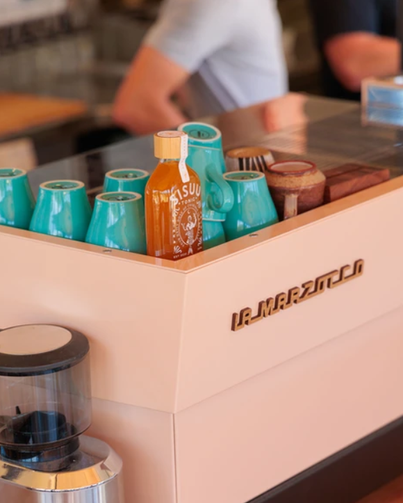 A countertop with teal cups, a bottle of Sriracha hot sauce, and a coffee grinder in a cafe setting.