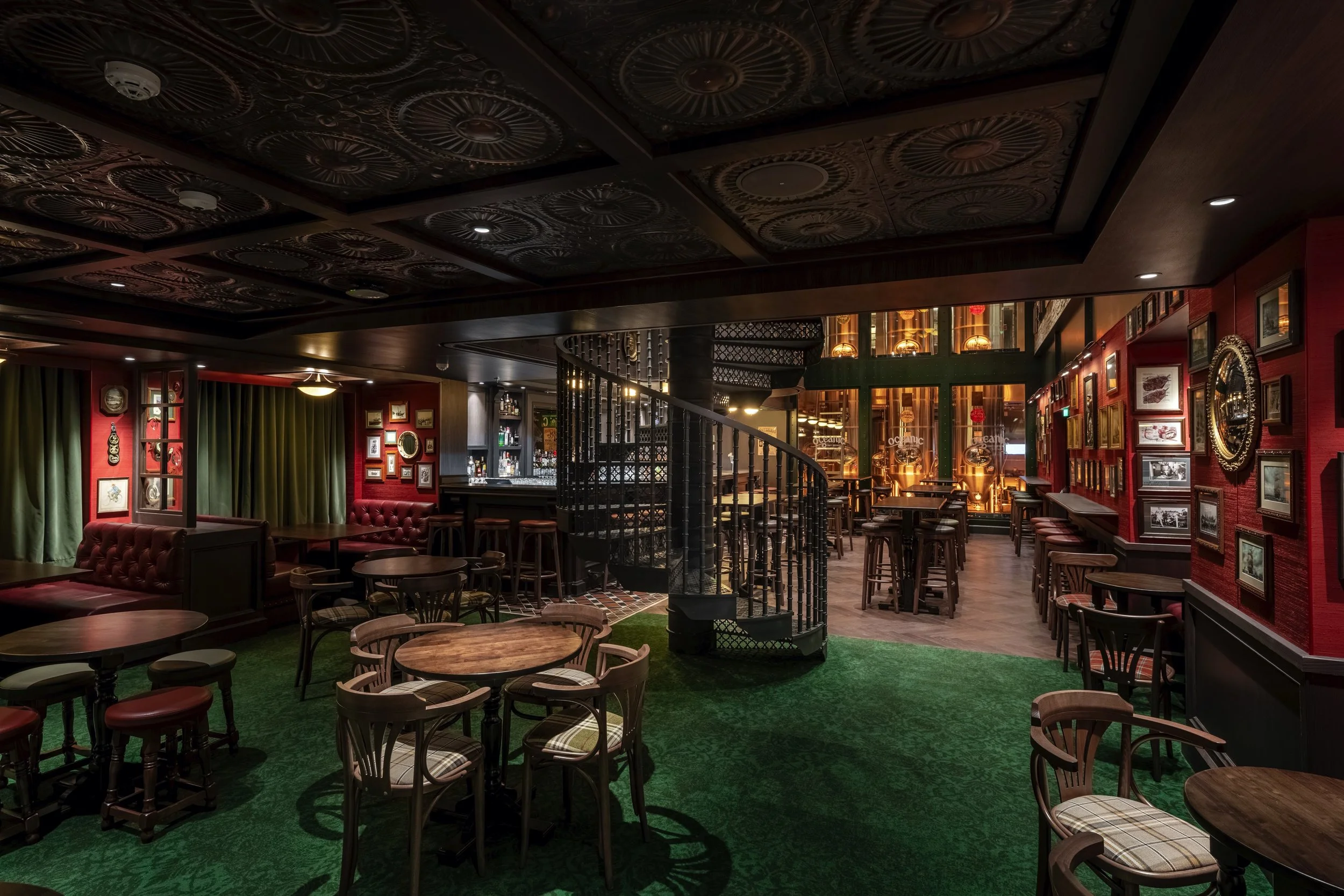 Interior of a cozy pub or bar with wooden tables and chairs, a spiral staircase at the center, framed pictures on red walls, green carpet, and a well-lit bar area in the background.