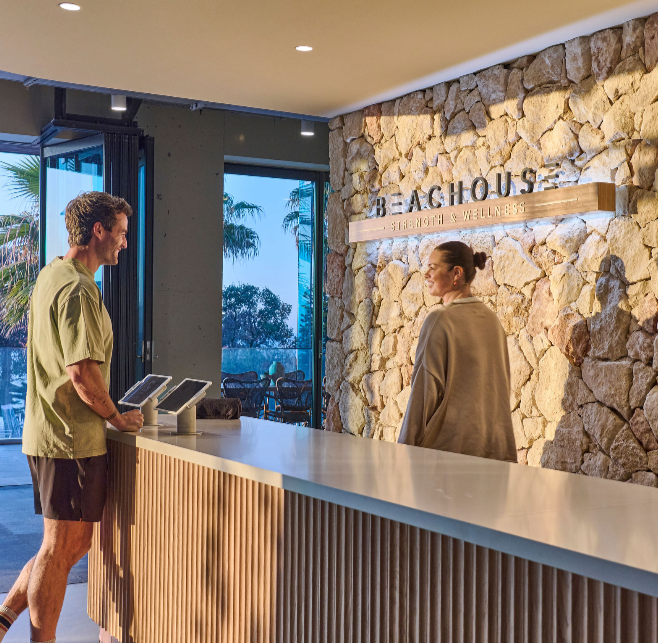 Man checking in at the reception desk of a wellness resort called Beachouse, with a stone wall behind the receptionist and large windows showing outdoor palm trees.