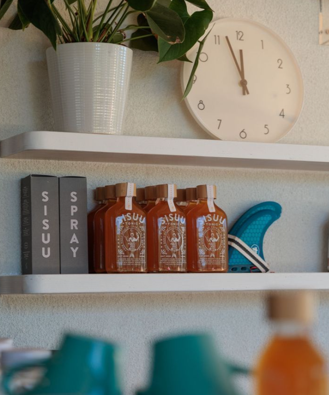 Wall shelf with potted plant, round wall clock, boxes labeled 'SISU' and 'SPRAY', three bottles of Sisu tonic, and a blue skateboard.