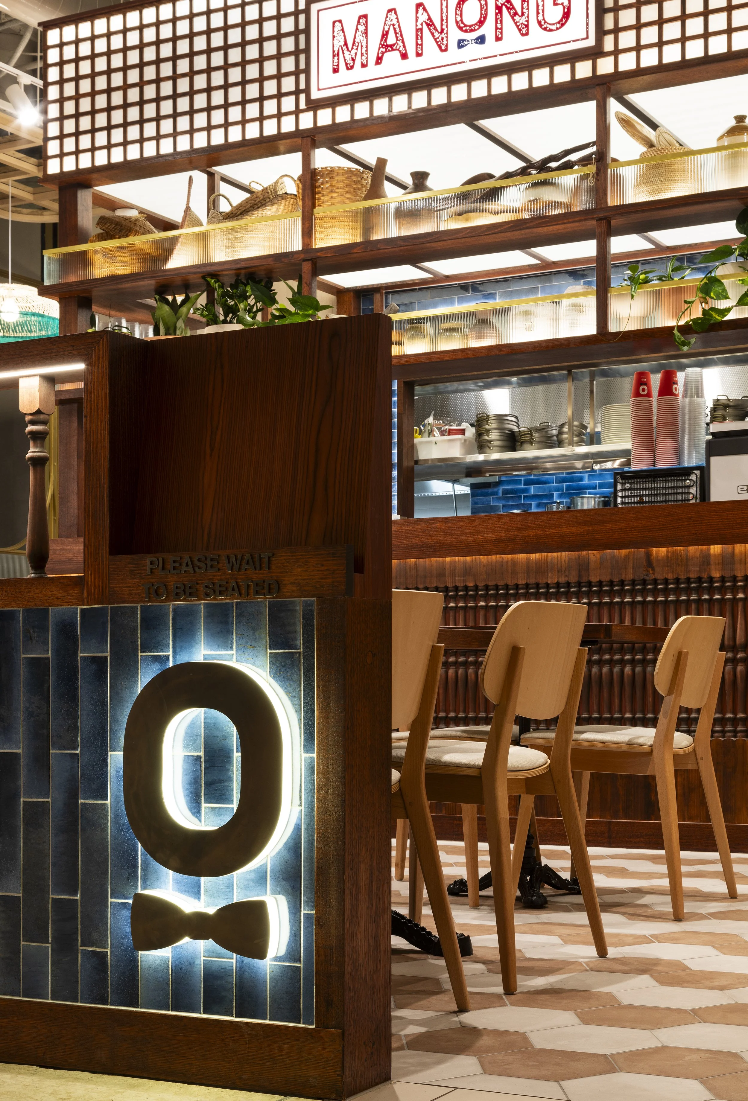Interior of a restaurant with a visible sign that reads 'MANNING'. There are light brown chairs grouped together, a wooden partition with blue tiles, and open kitchen shelves with dishes and containers. The floor has a hexagonal tile pattern.