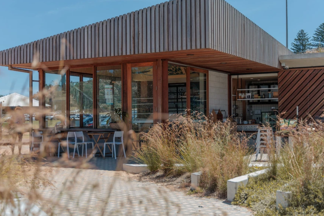 Modern building with large glass windows, wooden accents, and outdoor seating surrounded by dry grass and plants under a blue sky.