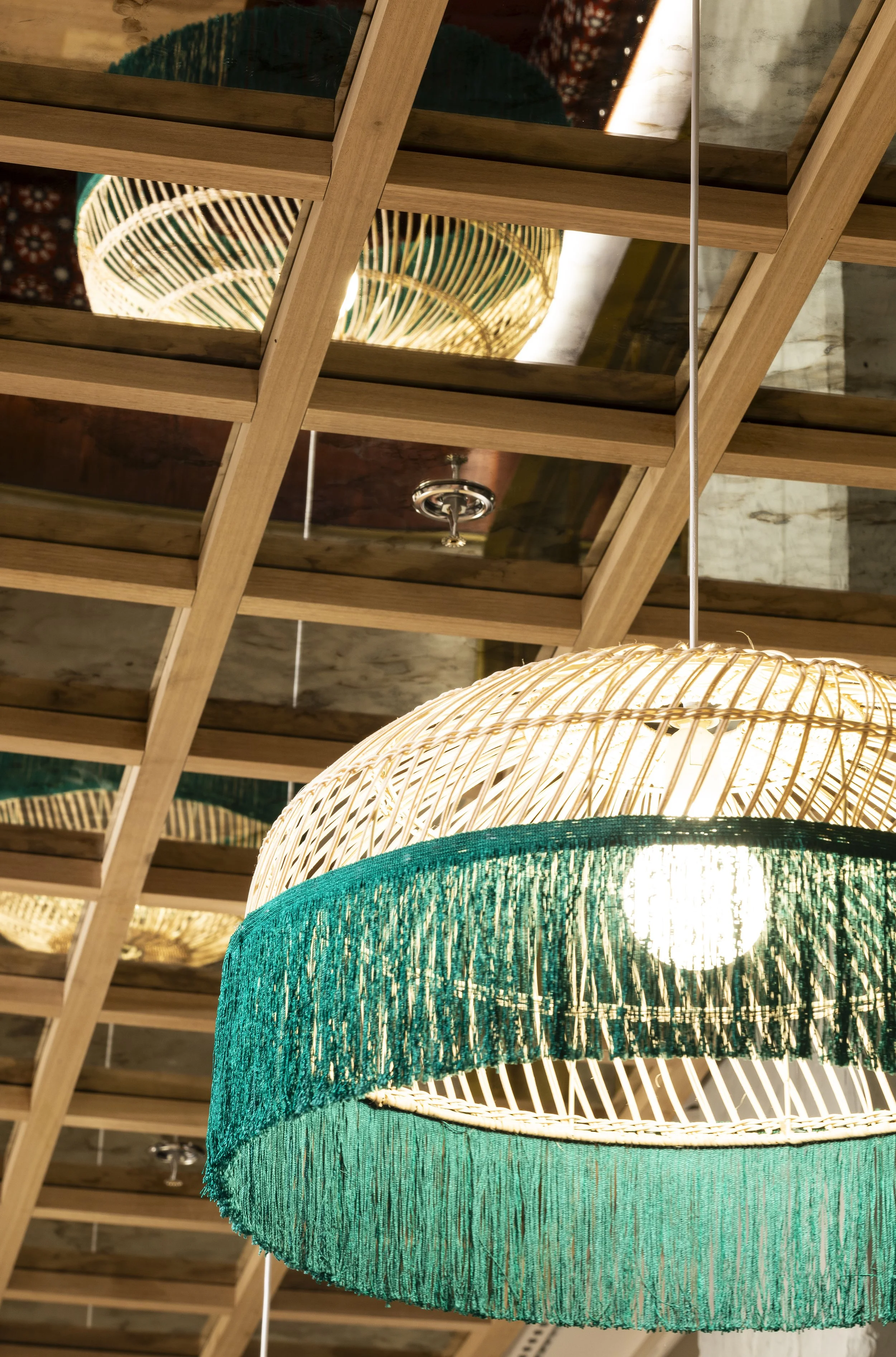 Close-up of a woven pendant light with a green fabric fringe, hanging from a wooden ceiling with mirrored panels.