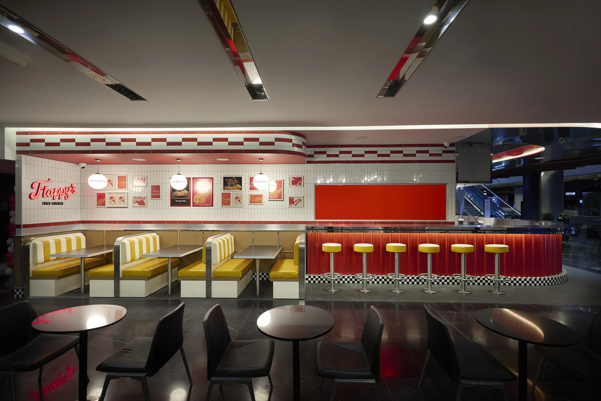 Interior of a retro-style fast food restaurant with yellow booths, black tables, and a red counter with yellow stools, featuring checkered black and white tiles and framed pictures on the wall.