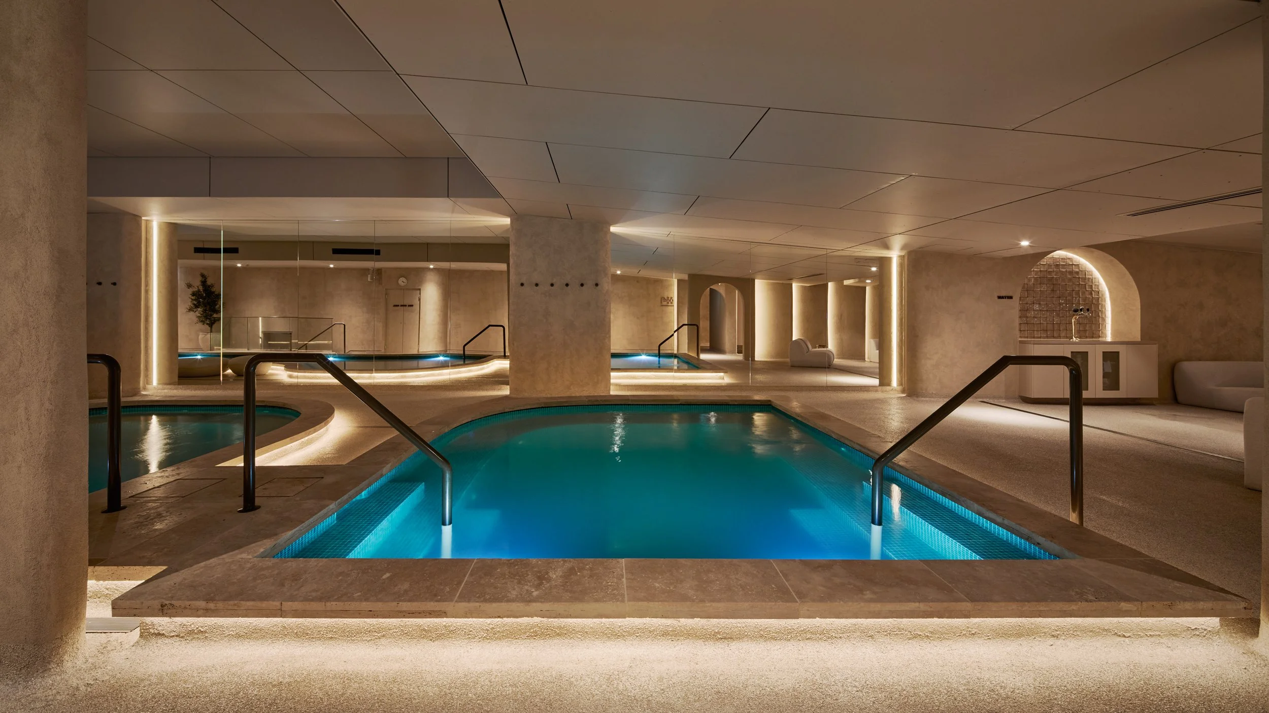 Indoor swimming pool area in a modern, minimalist spa with soft lighting, beige walls, and seating areas.