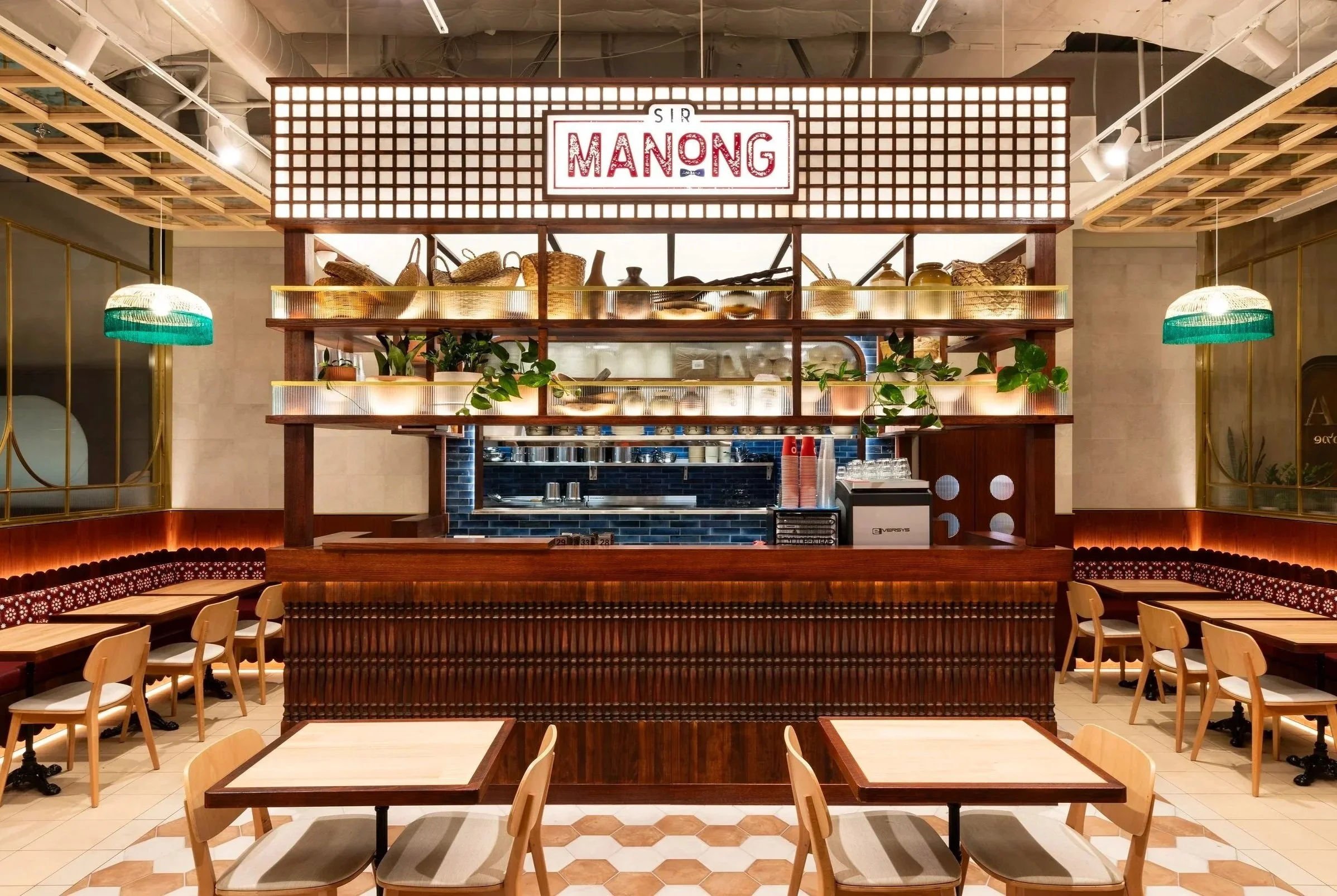 Interior of a restaurant with a central bar area, seating on both sides, and decorative hanging lights. The sign reads 'Sir Manong' above the bar.