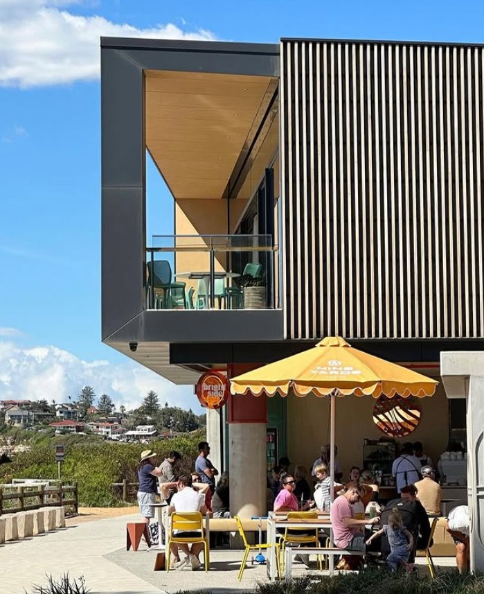 A modern building with a balcony on the upper floor and a café on the ground level. The café has outdoor seating under a yellow umbrella, with people dining outside on a sunny day.