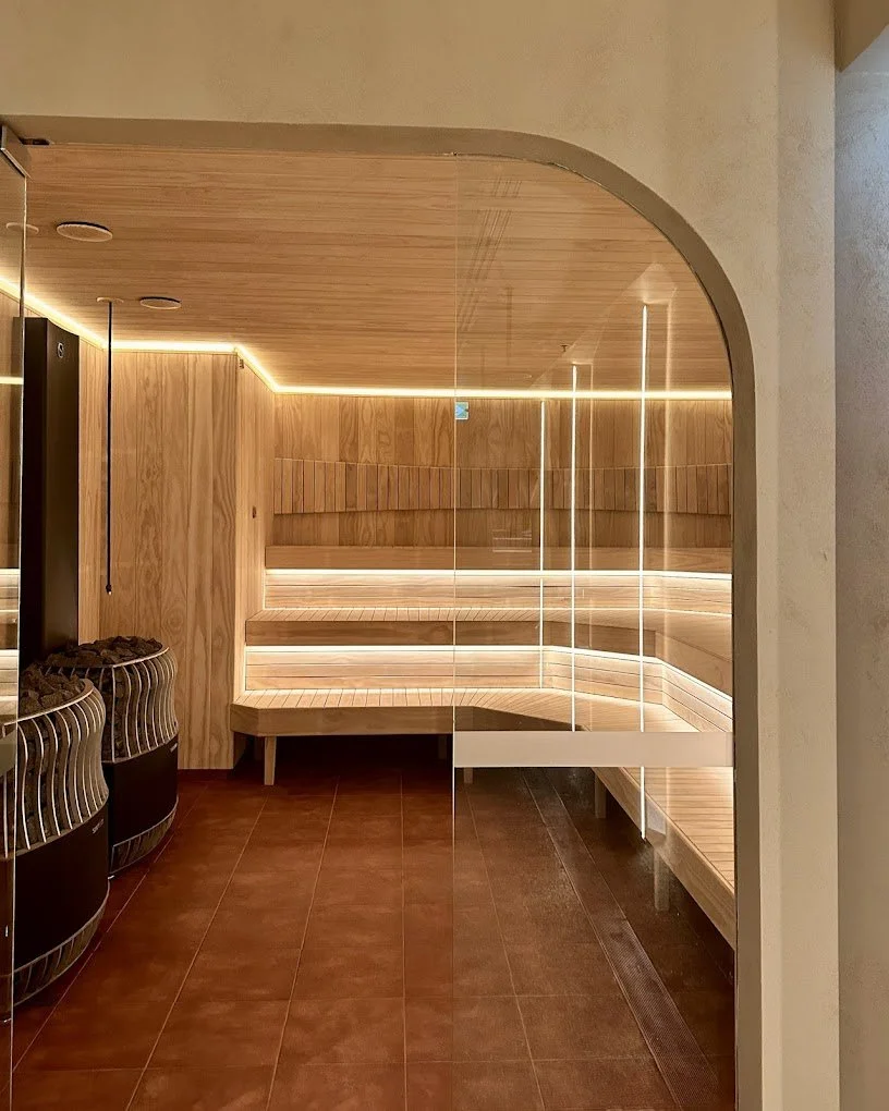 A modern, wooden sauna room with bench seating illuminated by LED strip lights, viewed through a glass door with a curved top.