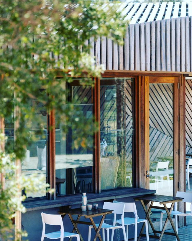 Outdoor patio seating area with white chairs, small black tables, and large glass windows of a building with wooden accents.