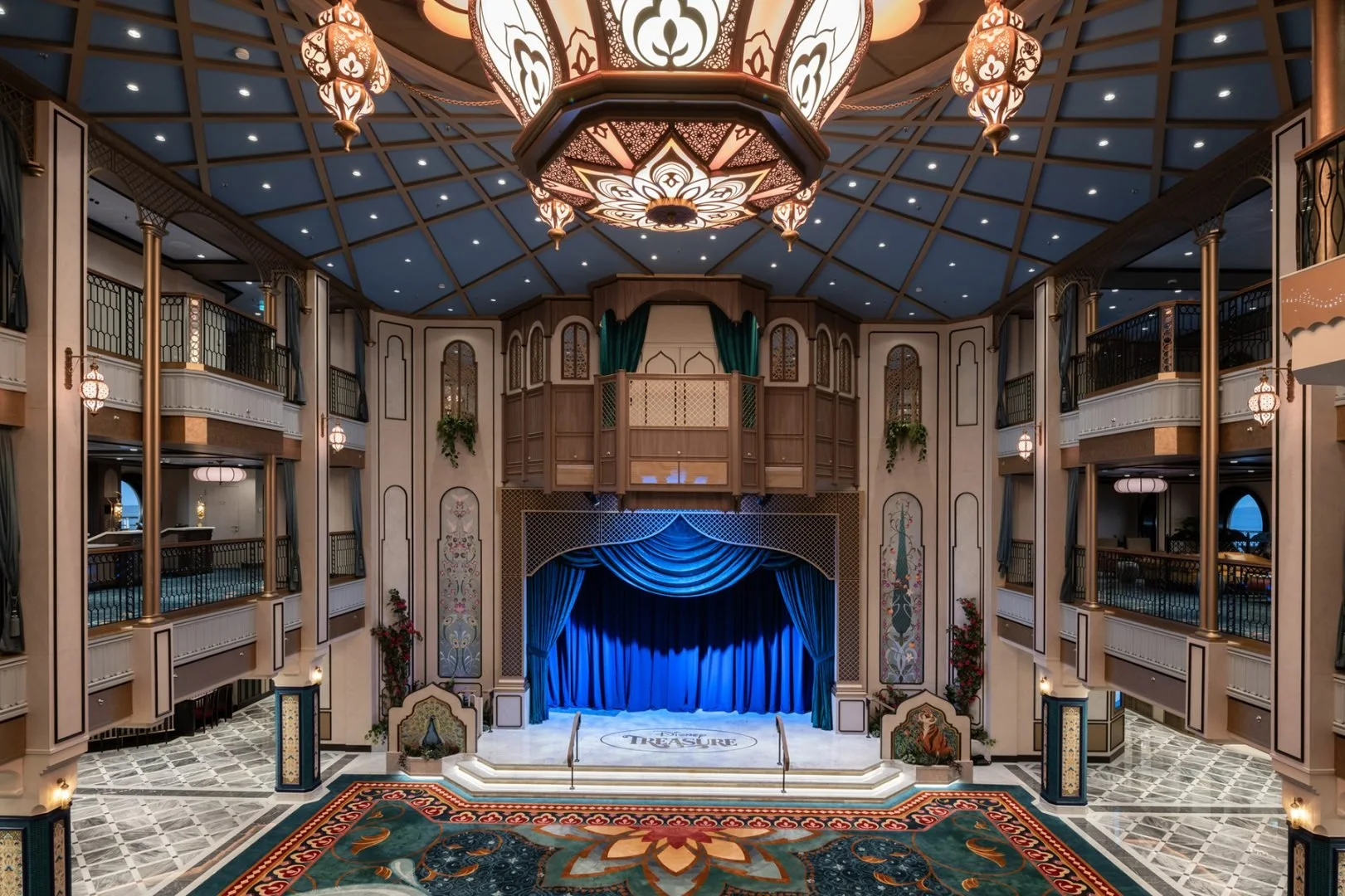 Interior of a grand theater with a stage framed by blue curtains, ornate walls, and a decorative ceiling with hanging lamps, viewed from the audience area.