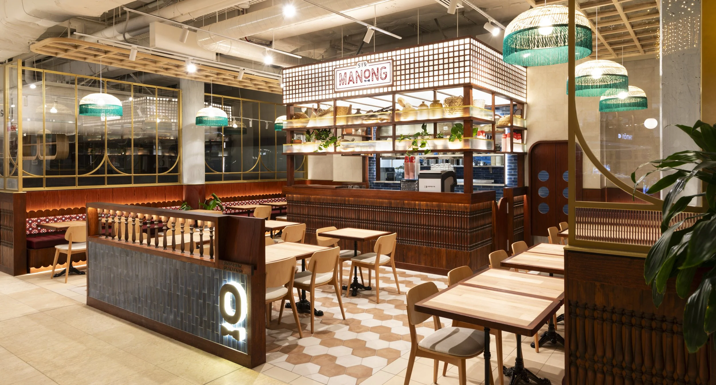 Interior of a modern restaurant or cafe with wooden tables and chairs, decorative hanging green and white pendant lights, a counter with a sign reading 'SIR MANONG,' and a glass partition with gold framing.