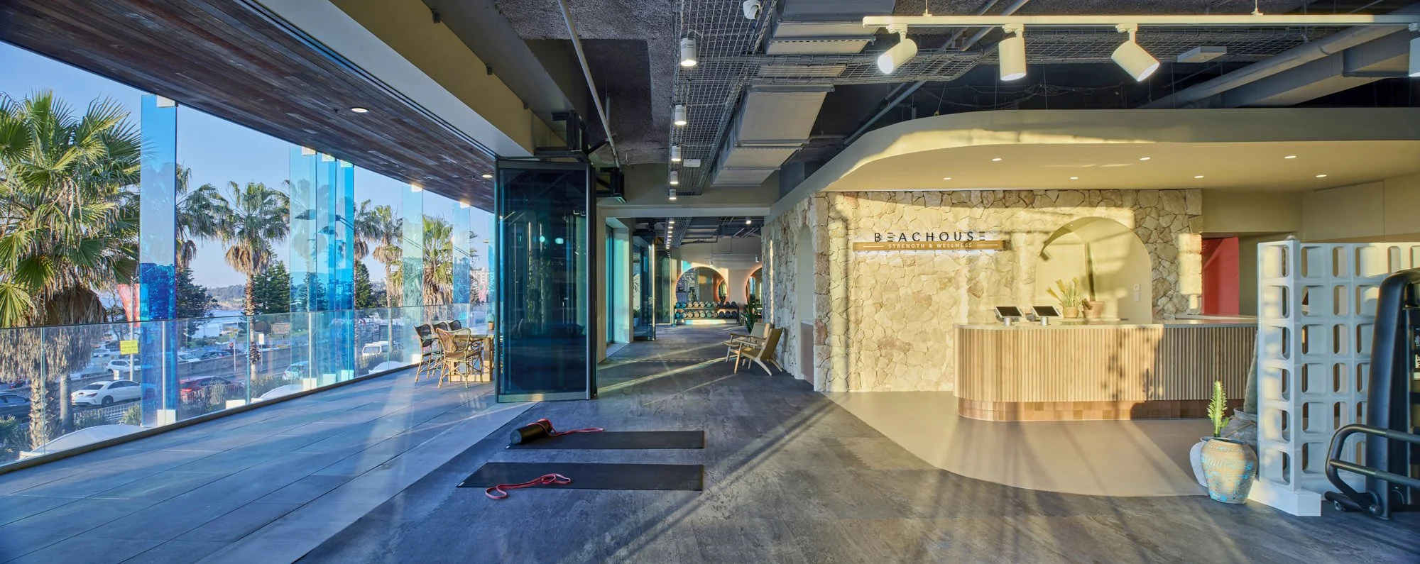 Interior of a fitness or wellness center with a reception desk, glass wall windows showing palm trees outside, yoga mats on the floor, and exercise equipment in the background.