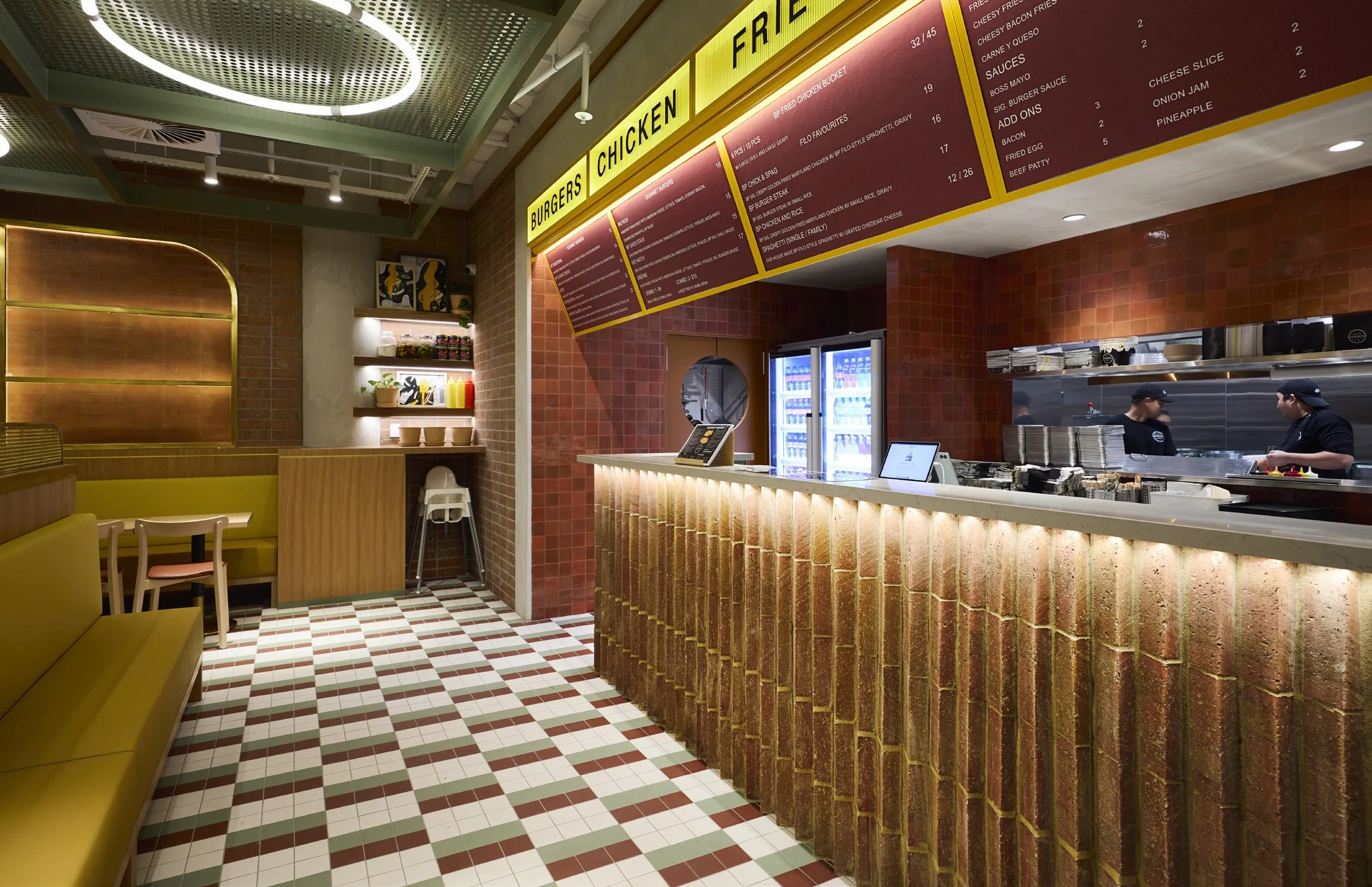 Interior of a fast food restaurant with a brick front counter, menu boards overhead, kitchen staff visible through a window, and yellow seating area to the left with tiled flooring.