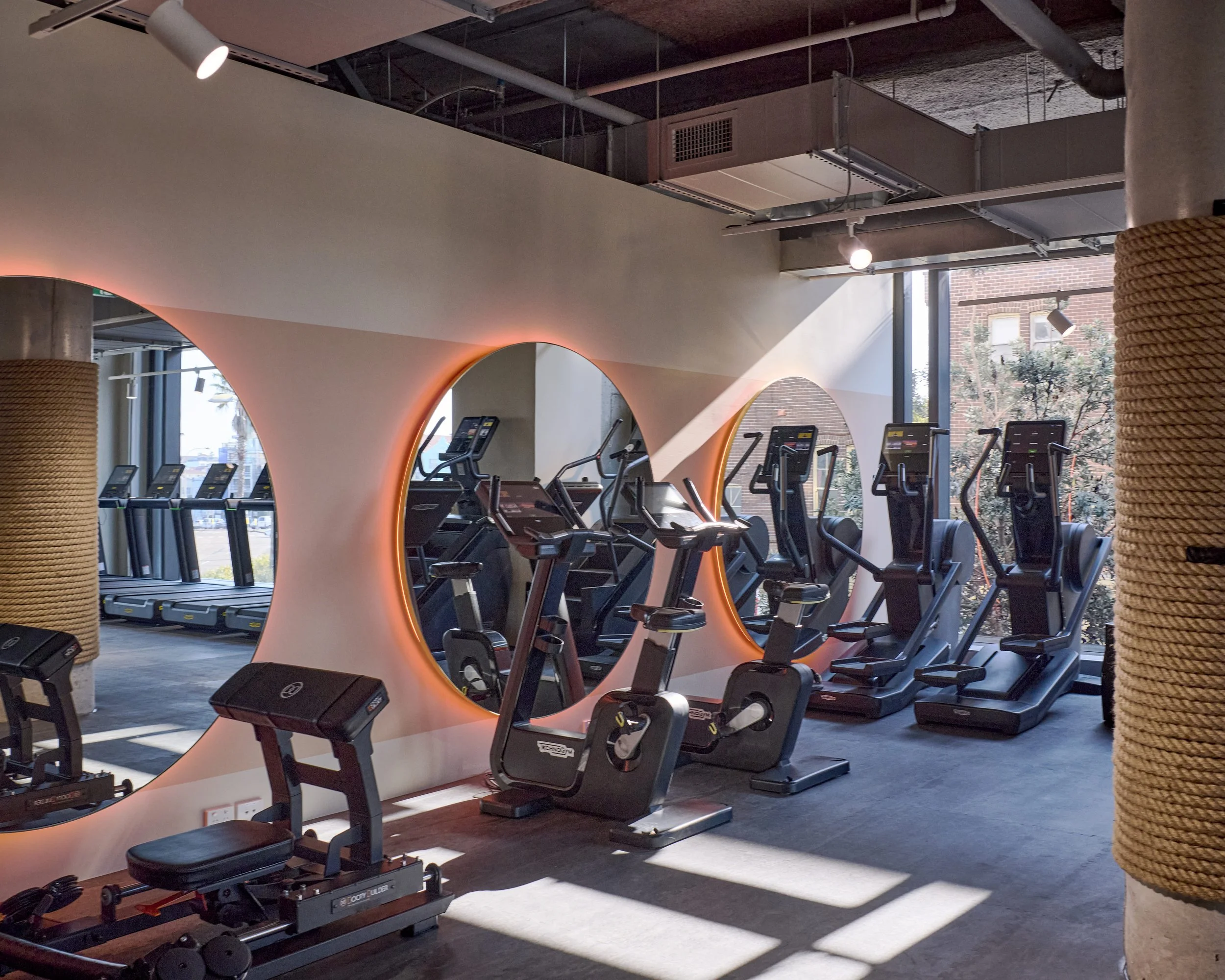 Modern gym with cardio equipment including treadmills and stationary bikes, illuminated by natural light from large windows, with decorative round mirrors and industrial-style ceiling.