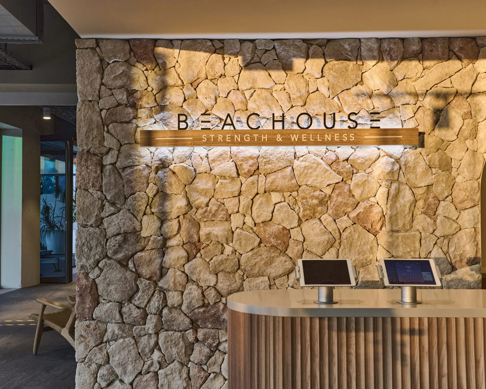 Reception area with a stone wall display reading 'Beachouse Strength & Wellness' and two digital tablets on a wooden counter.