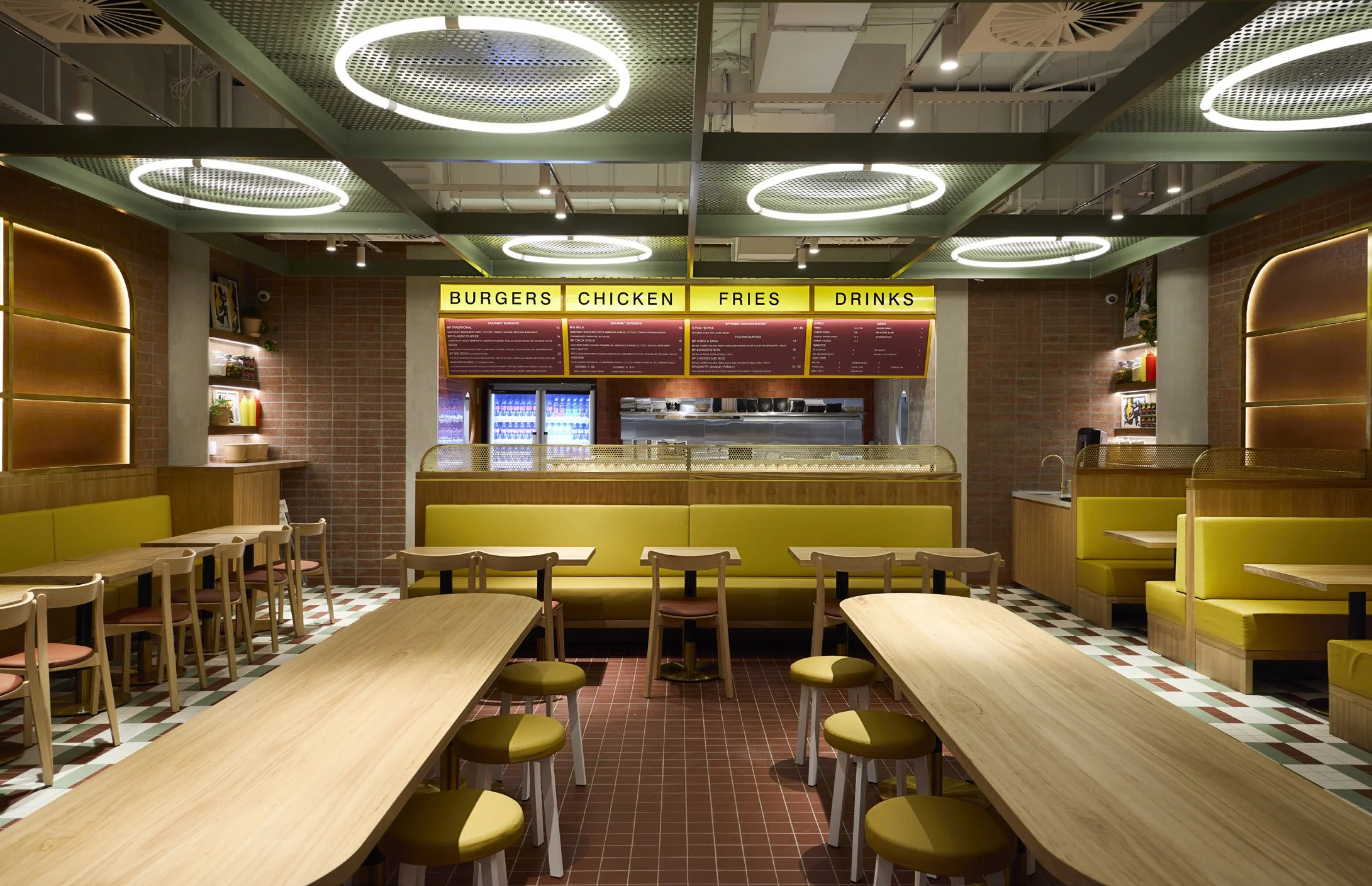 Empty fast-food restaurant interior with yellow booths, wooden tables, and chairs, menu boards overhead with categories like Burgers, Chicken, Fries, Drinks, brick walls, and modern lighting fixtures.