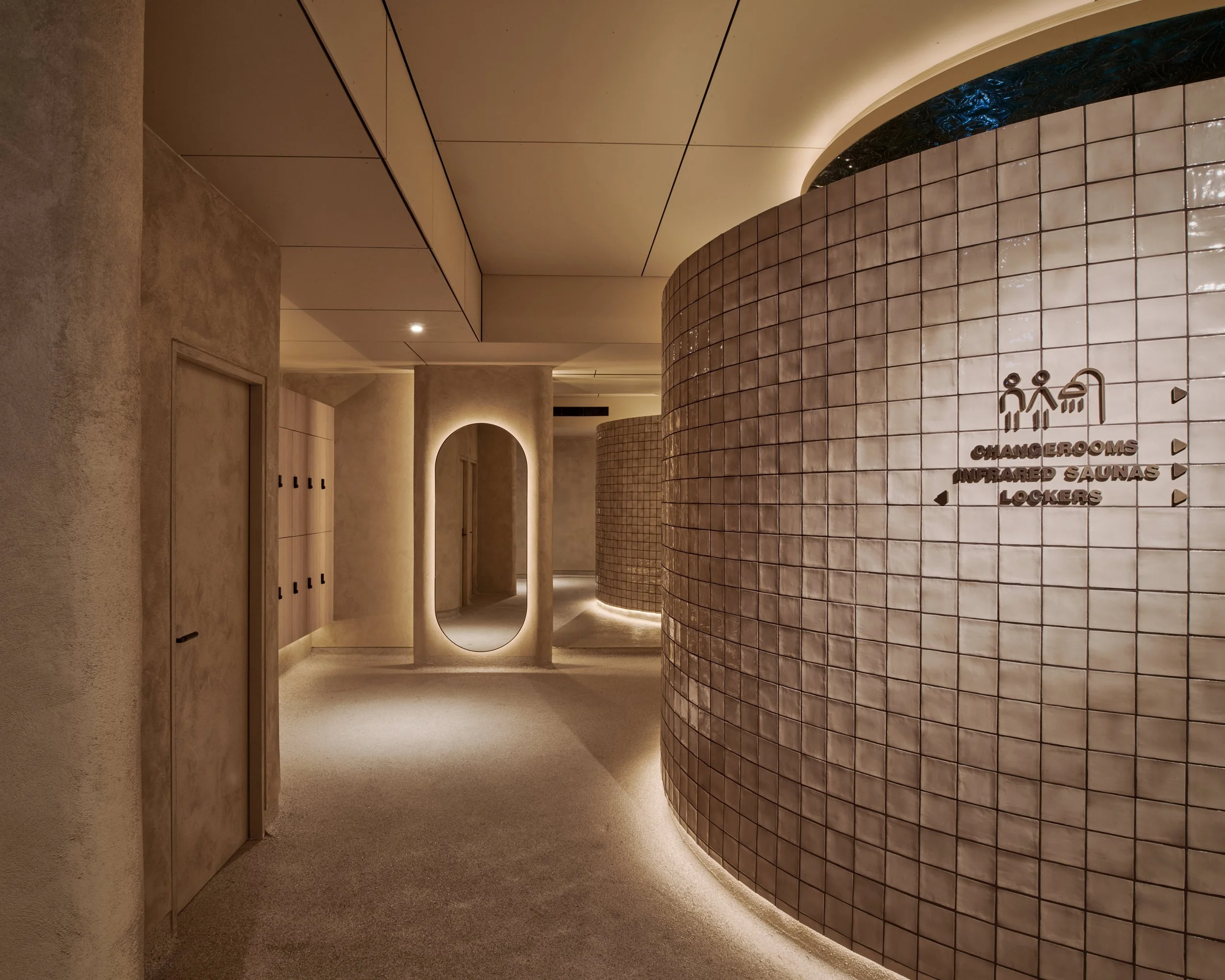 Modern spa or wellness center corridor with curved tiled walls, lockers on the left, a mirror with backlighting, and a sign indicating changing rooms, infrared saunas, and lockers.