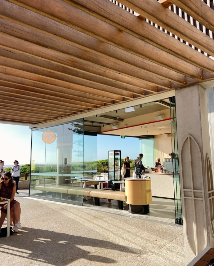 Interior of a modern café with a glass wall, customers ordering at the counter, and outdoor seating visible through the glass.