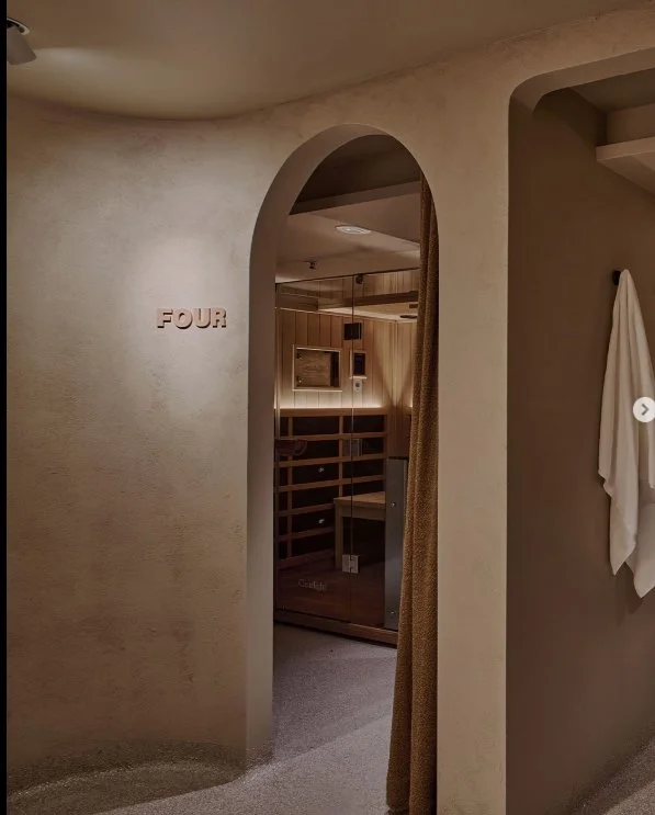 A beige hallway with an arched doorway leading into a room with wooden walls and furniture, a wall-mounted TV, and a towel hanging on the wall to the right. The word 'FOUR' is mounted on the left wall.