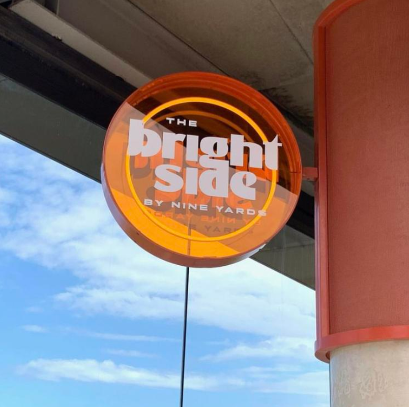 Sign for The Bright Side restaurant by Nine Yards, with orange neon lighting and white text, against a background of blue sky with clouds.
