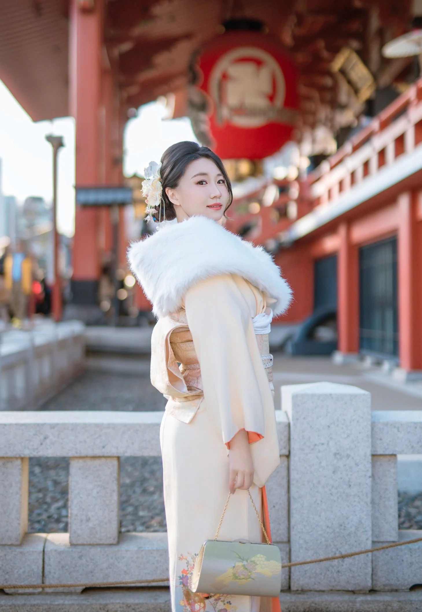 A young woman dressed in a traditional Japanese kimono with a white fur stole, standing outdoors near a Japanese temple or shrine. She has floral hair accessories and holds a small, elegant purse.