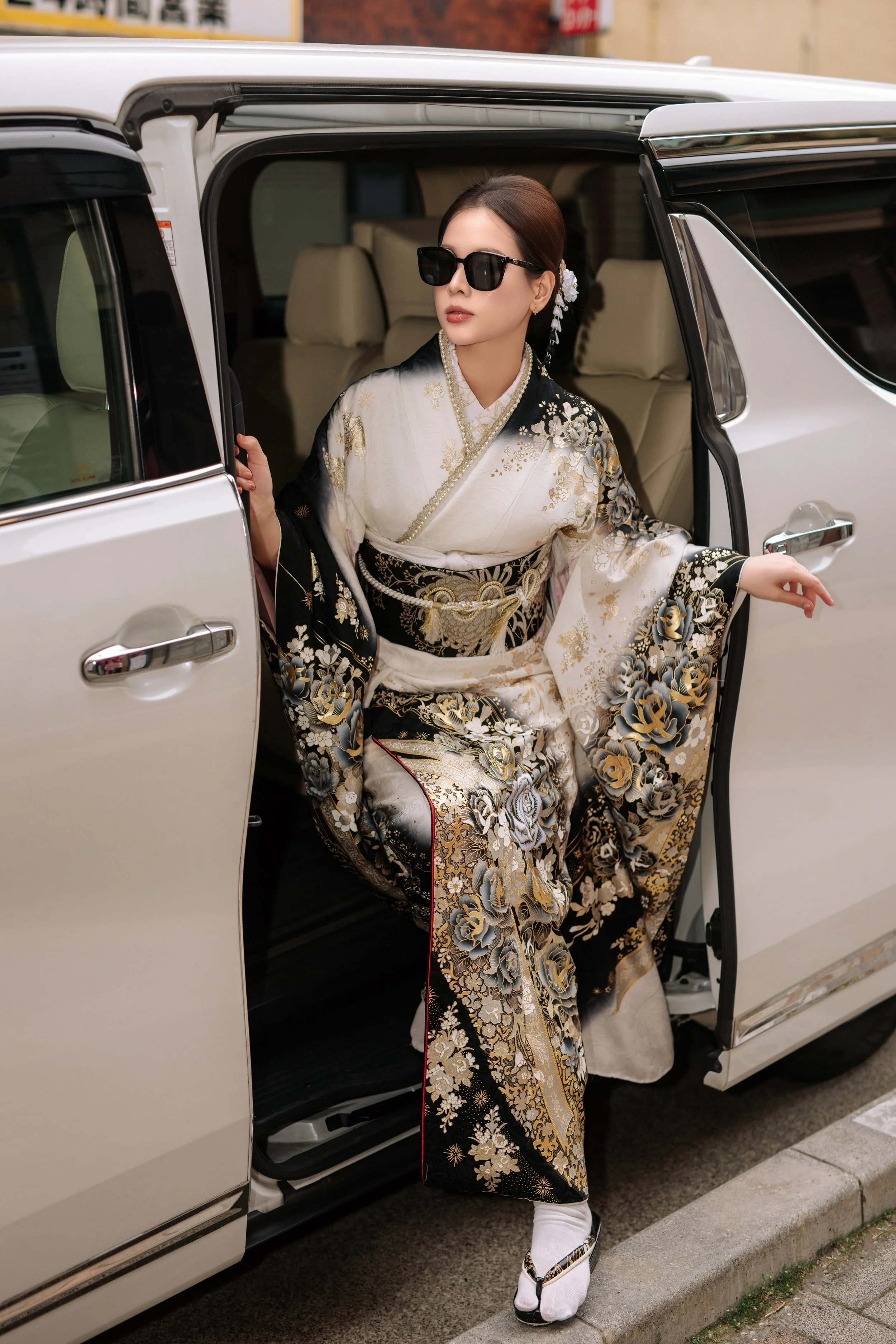 A woman dressed in an elegant black and white kimono with gold and floral patterns, wearing large black sunglasses, and sitting in a white luxury car on a city street.