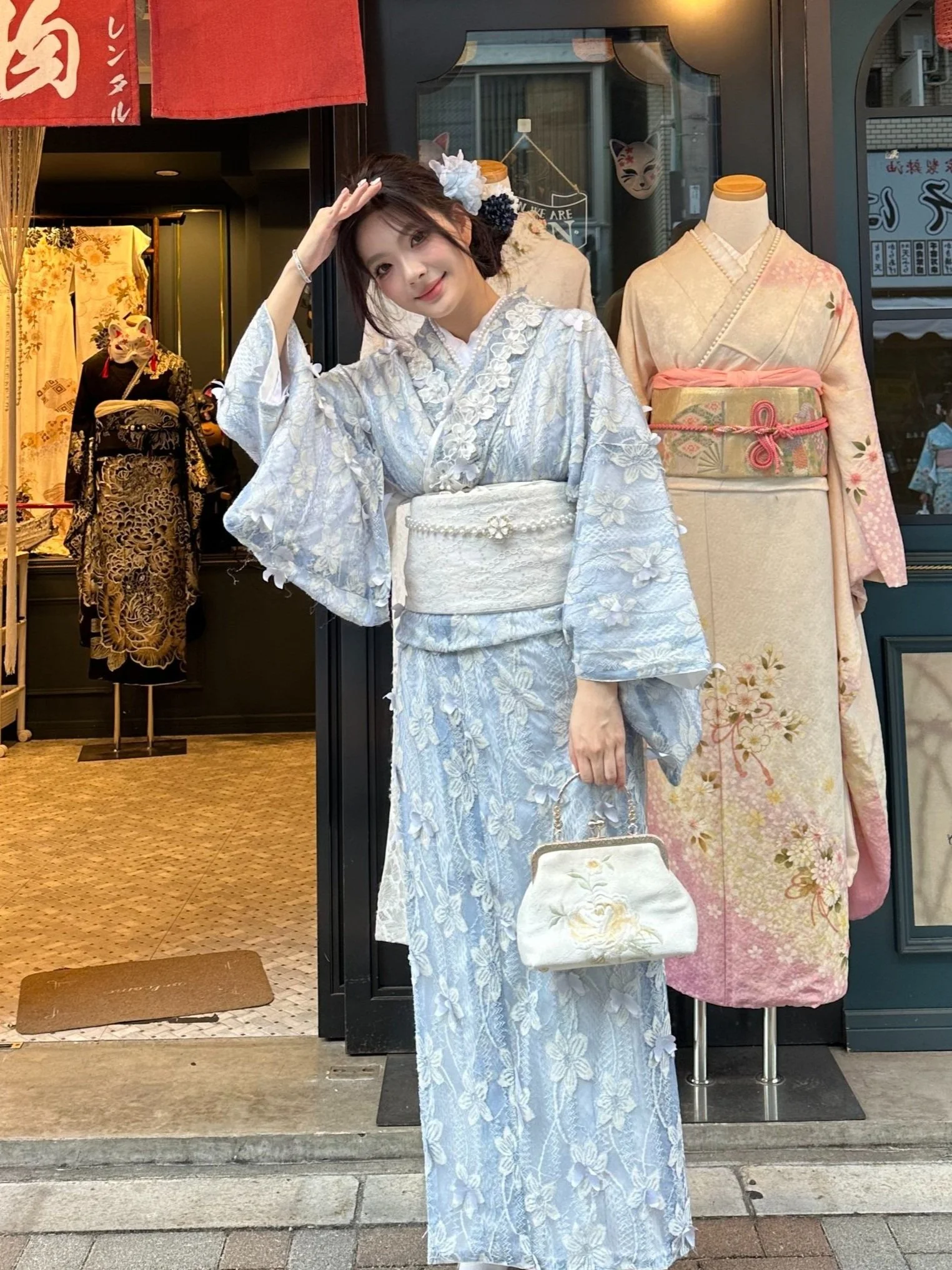 Young woman in a white and silver floral kimono, standing in front of a store with traditional mannequins wearing kimonos. The woman is holding a white embroidered handbag and has flowers in her hair, smiling and saluting.
